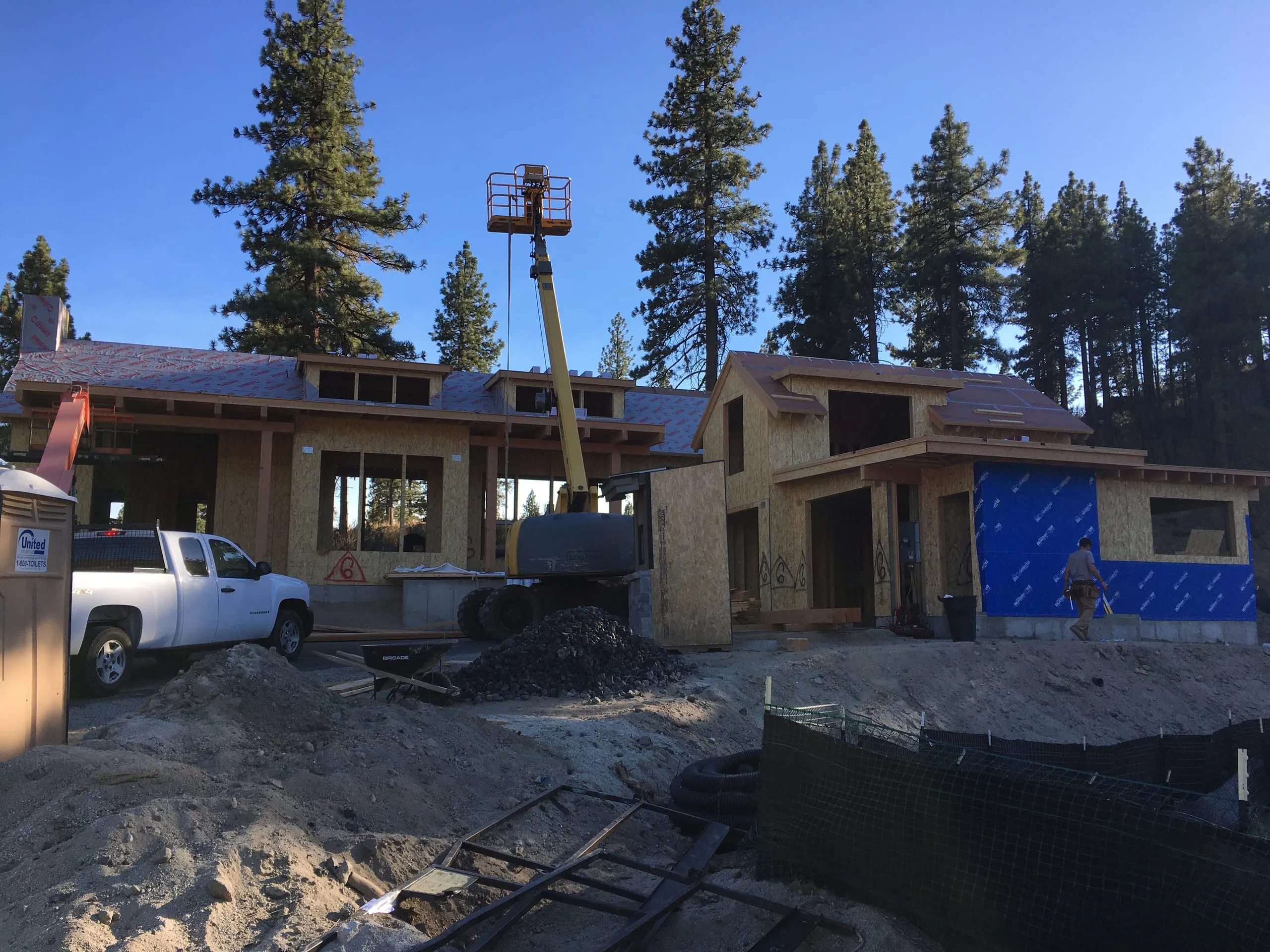 Construction photo: wood-framed split-level house with blue weatherproofing, a lift and a pickup truck among pines