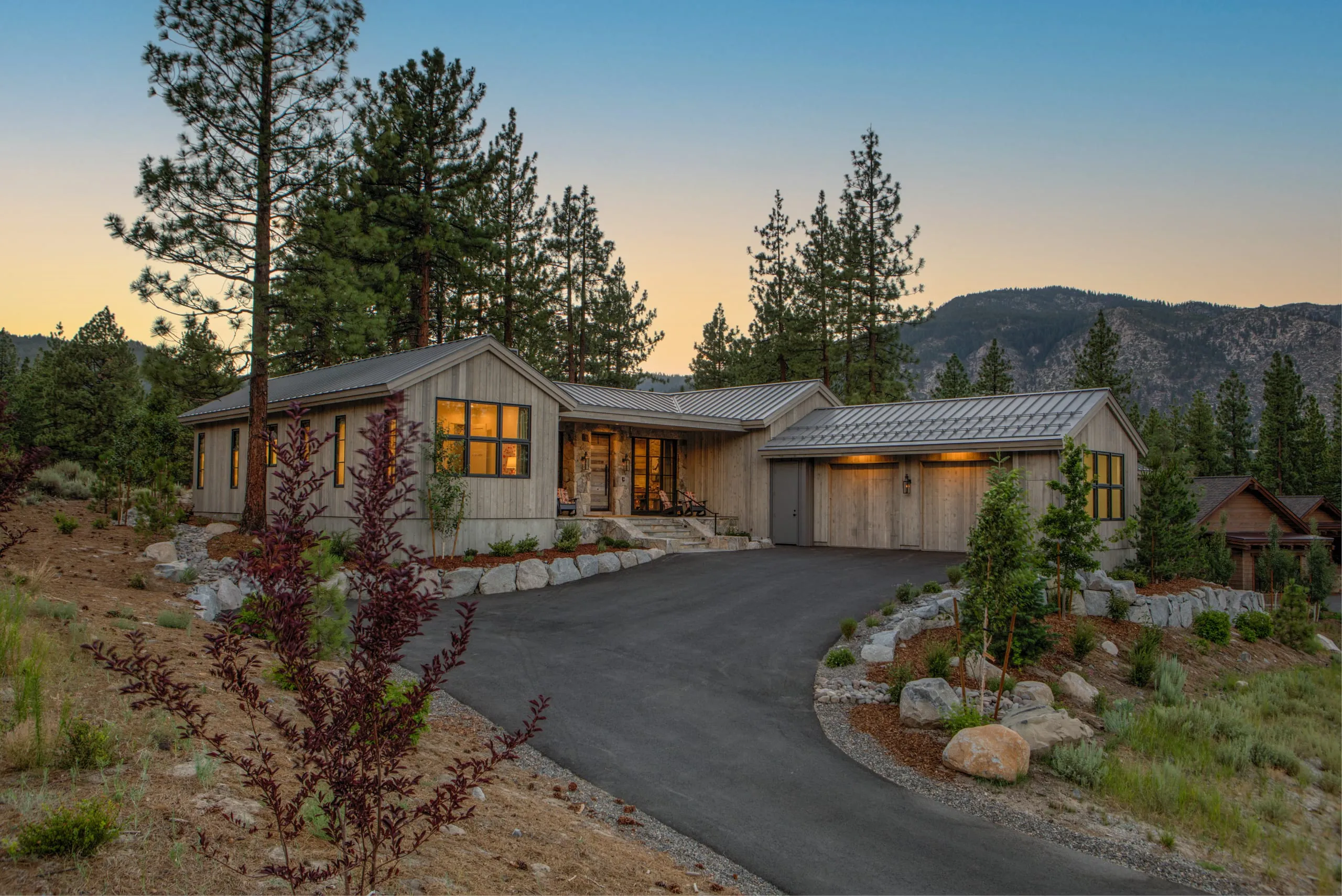 Modern farmhouse front exterior at dusk with vertical wood siding, metal roofs, garage, stone-lined driveway and mountain behind