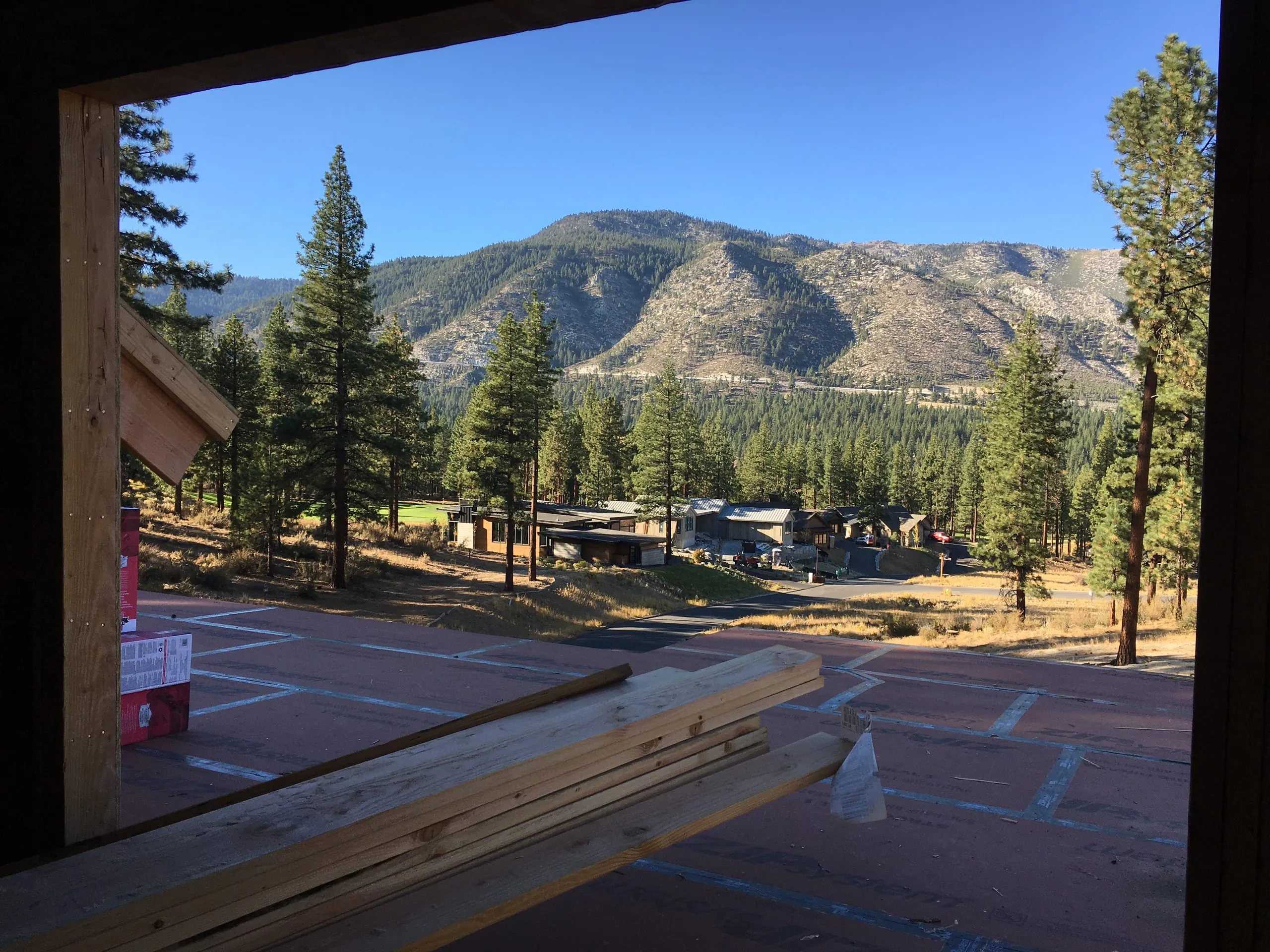 Construction photo: view from a framed opening across the slab to pine trees, nearby homes and a mountain slope