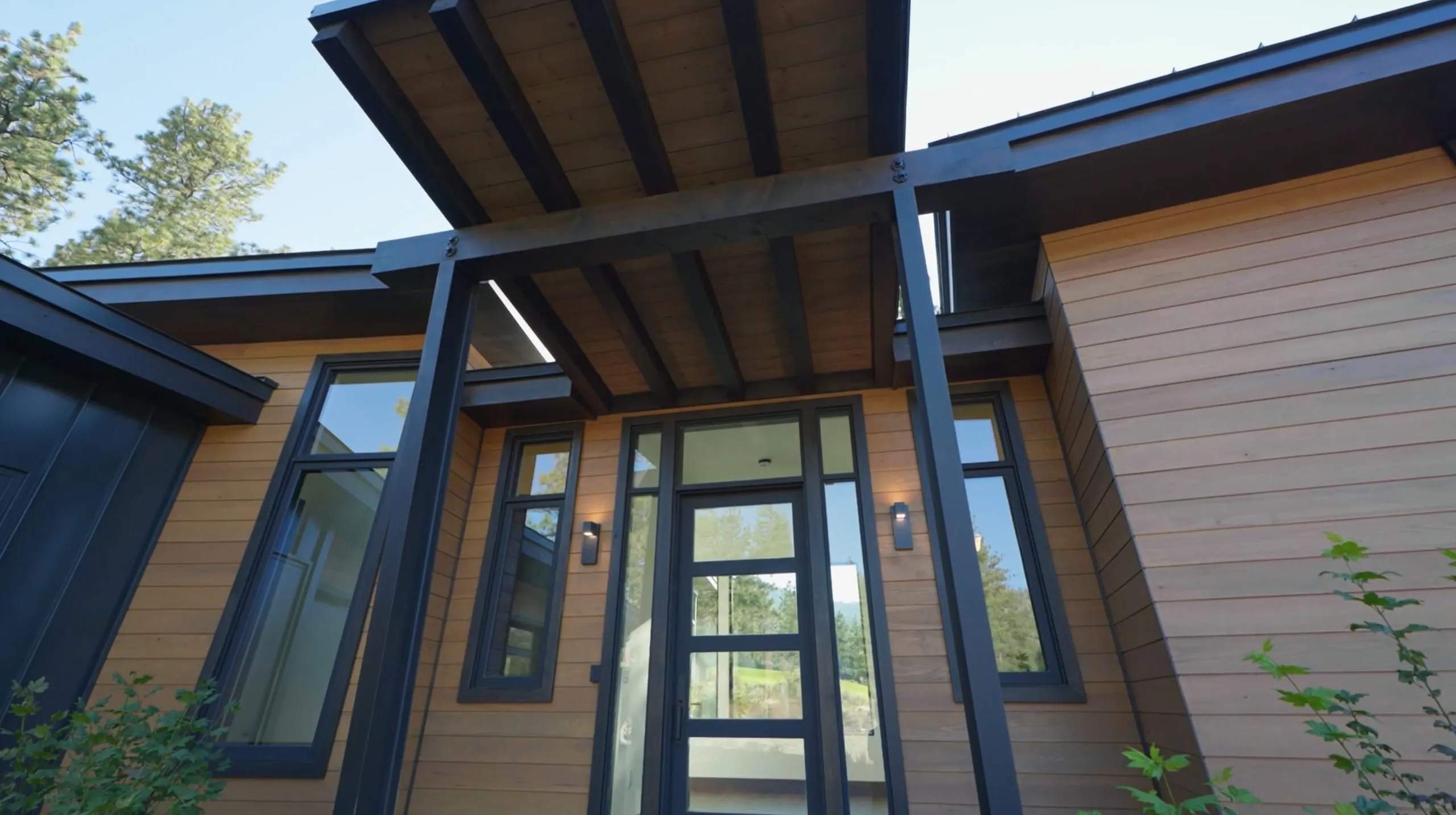 Entry with a steel-framed canopy over a glass front door, flanked by tall windows and warm horizontal wood siding