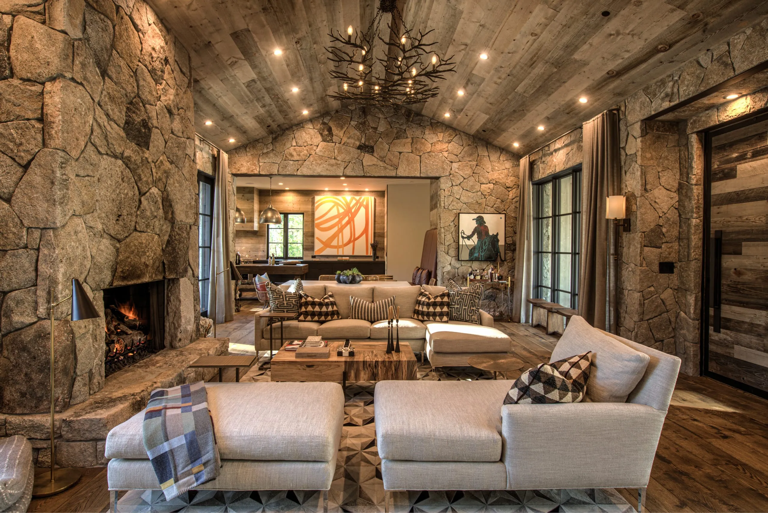 Vaulted living room with stone fireplace and walls, wood plank ceiling, antler chandelier and open view to the kitchen