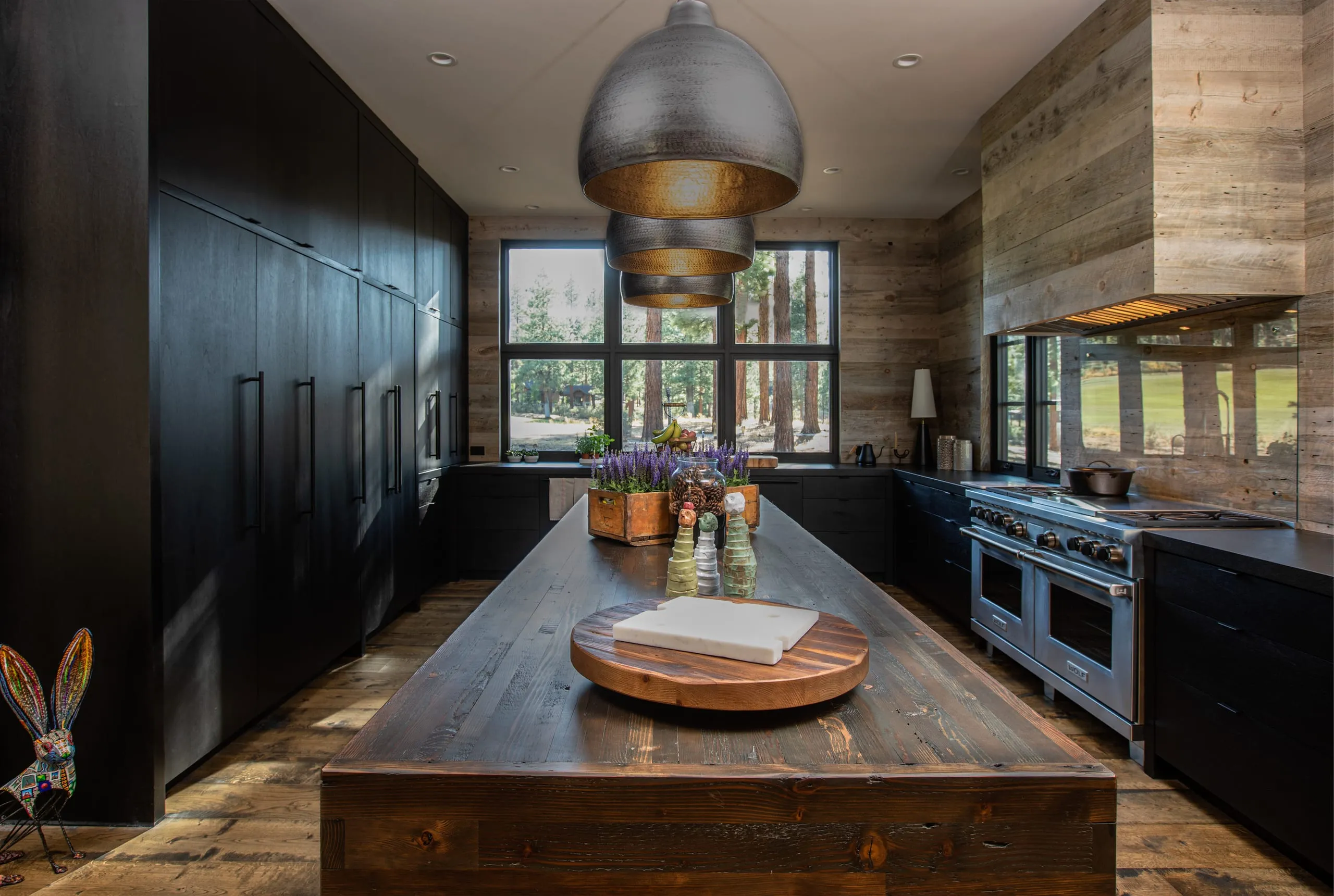 Kitchen with dark cabinetry, long reclaimed-wood island, pendant lights, range with wood hood and a large window to the trees