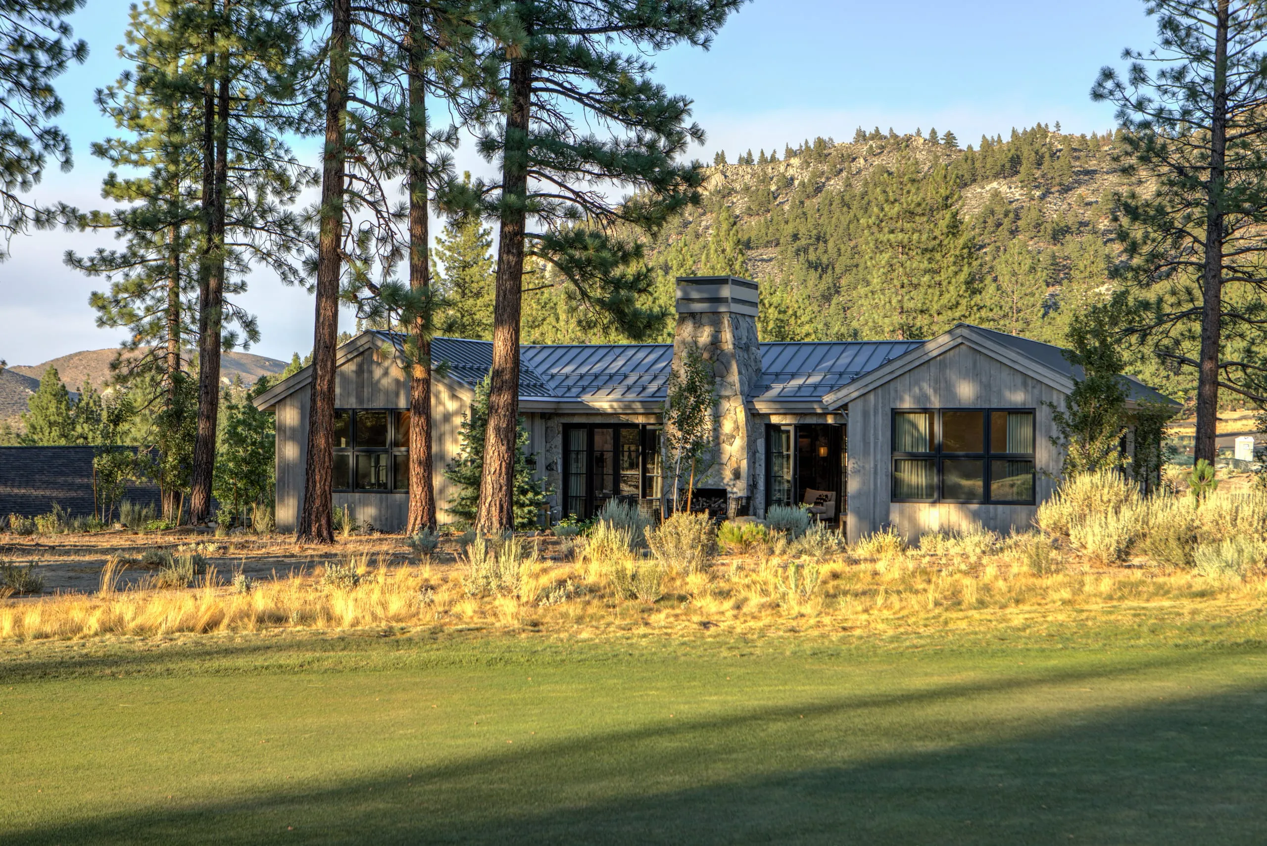 Rear exterior of a one-story house with gabled metal roof, stone chimney and gray wood siding, seen across a golf fairway