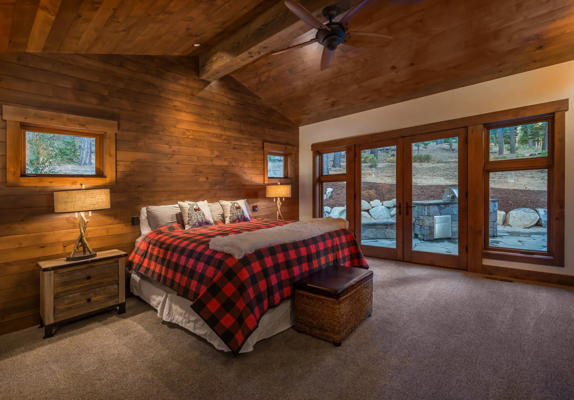 Bedroom with knotty wood walls and vaulted wood ceiling, red plaid bed and French doors opening to a stone patio