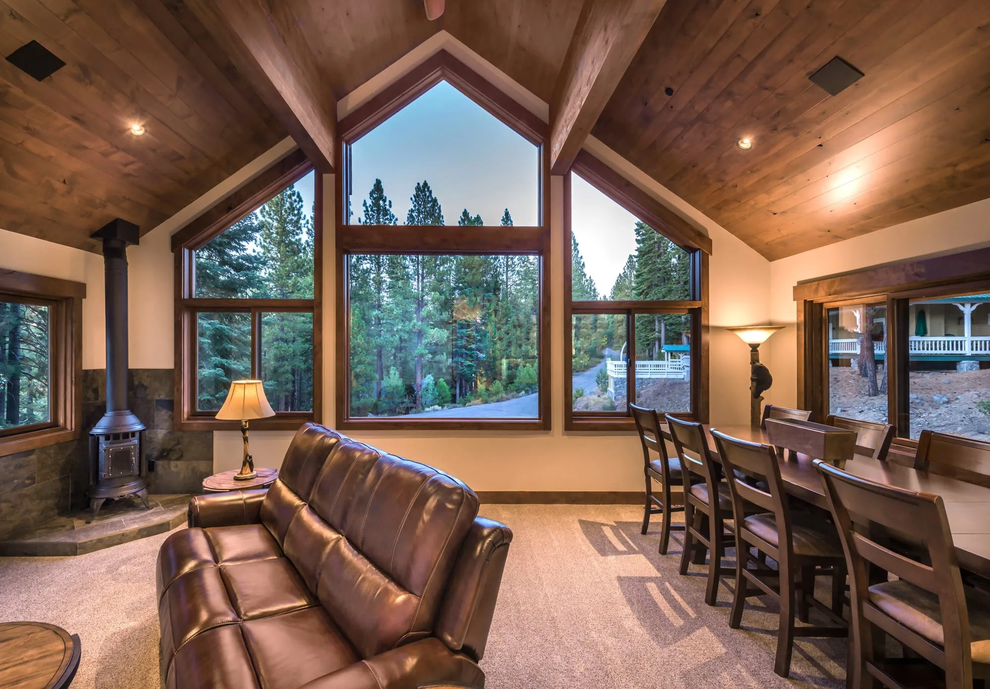 Living area with vaulted wood ceiling, tall peaked windows onto pine forest, a wood stove, leather sofa and dining table