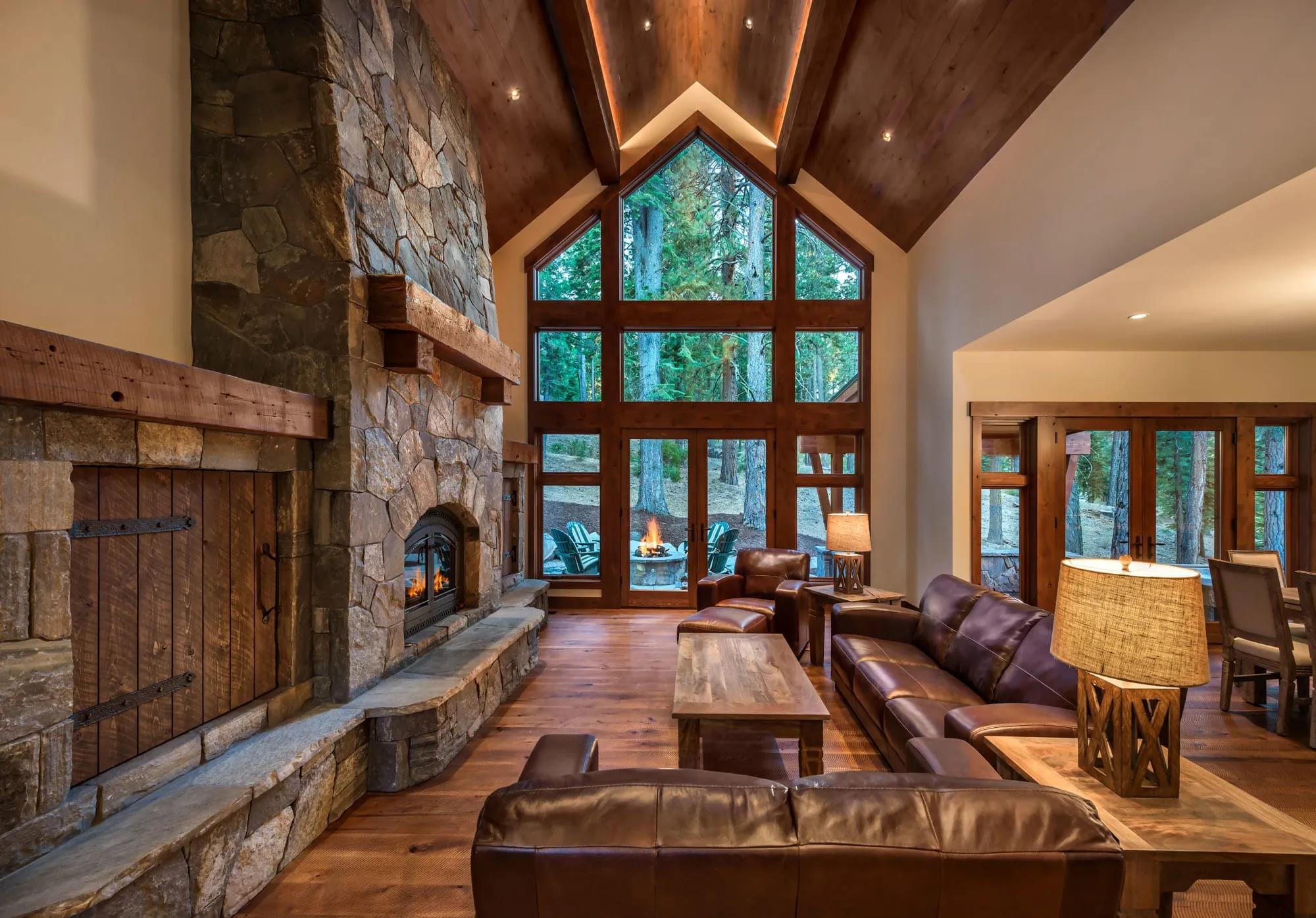 Living room with a tall stone fireplace, rustic wood doors, vaulted wood ceiling, peaked window wall and leather sofas