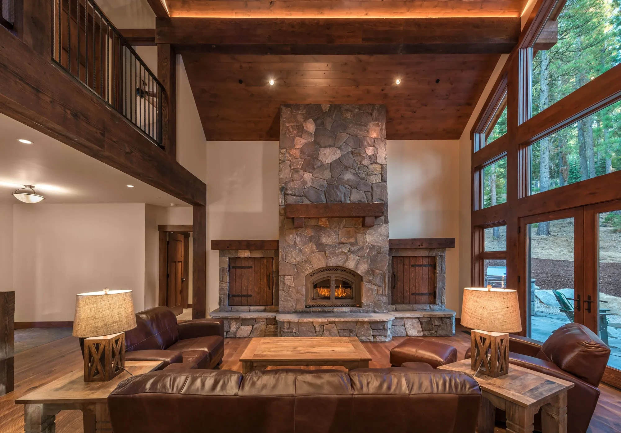 Living room with a central stone fireplace flanked by wood doors, timber ceiling beams, loft railing and a window wall