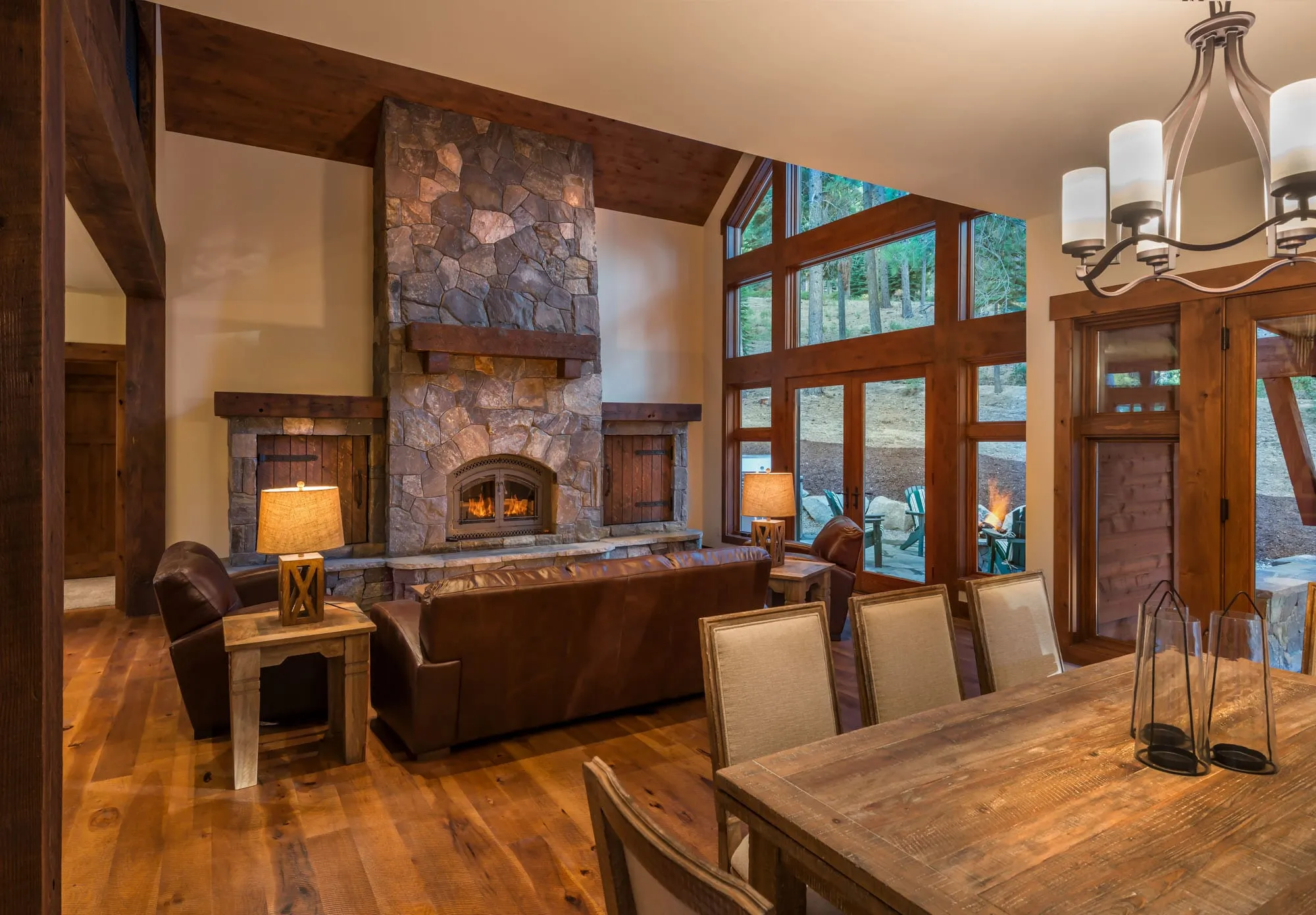 Great room with stone fireplace, timber posts, wood floors, leather seating, a dining table and tall windows to a patio
