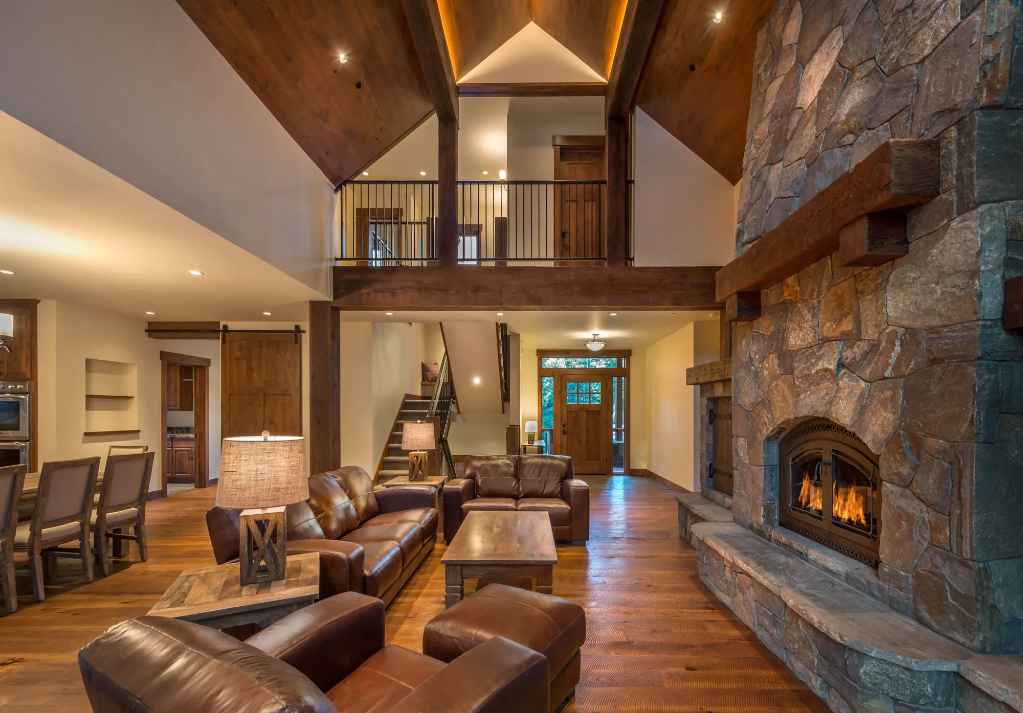 Open living room with timber beams, vaulted wood ceiling, loft railing, stone fireplace and an entry stair beyond