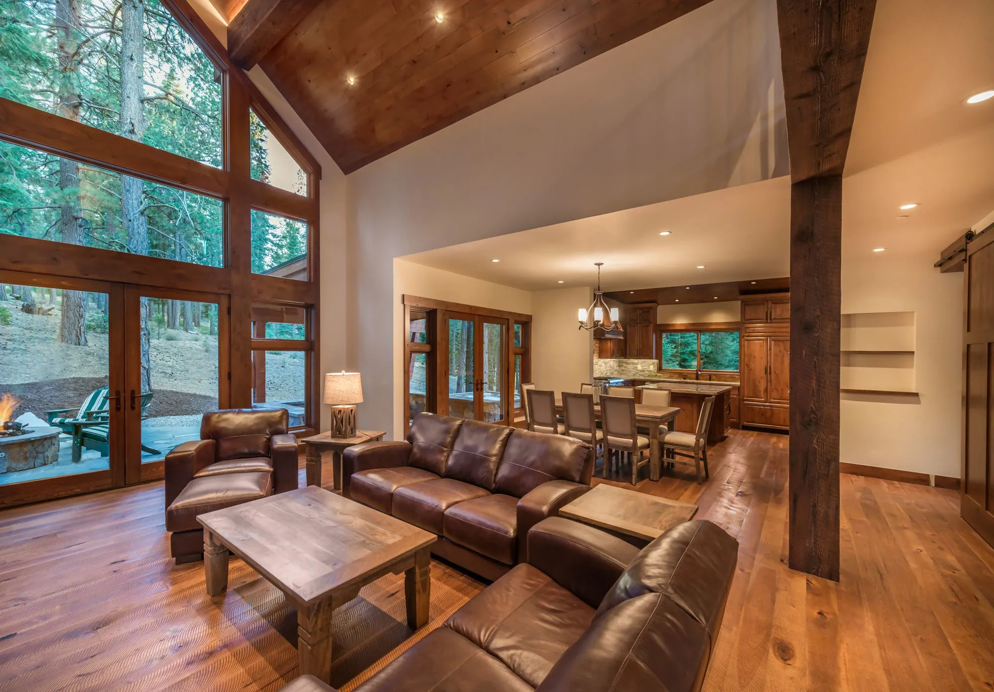 Vaulted living room with a tall window wall, timber post, leather sofas and an open dining area and kitchen beyond