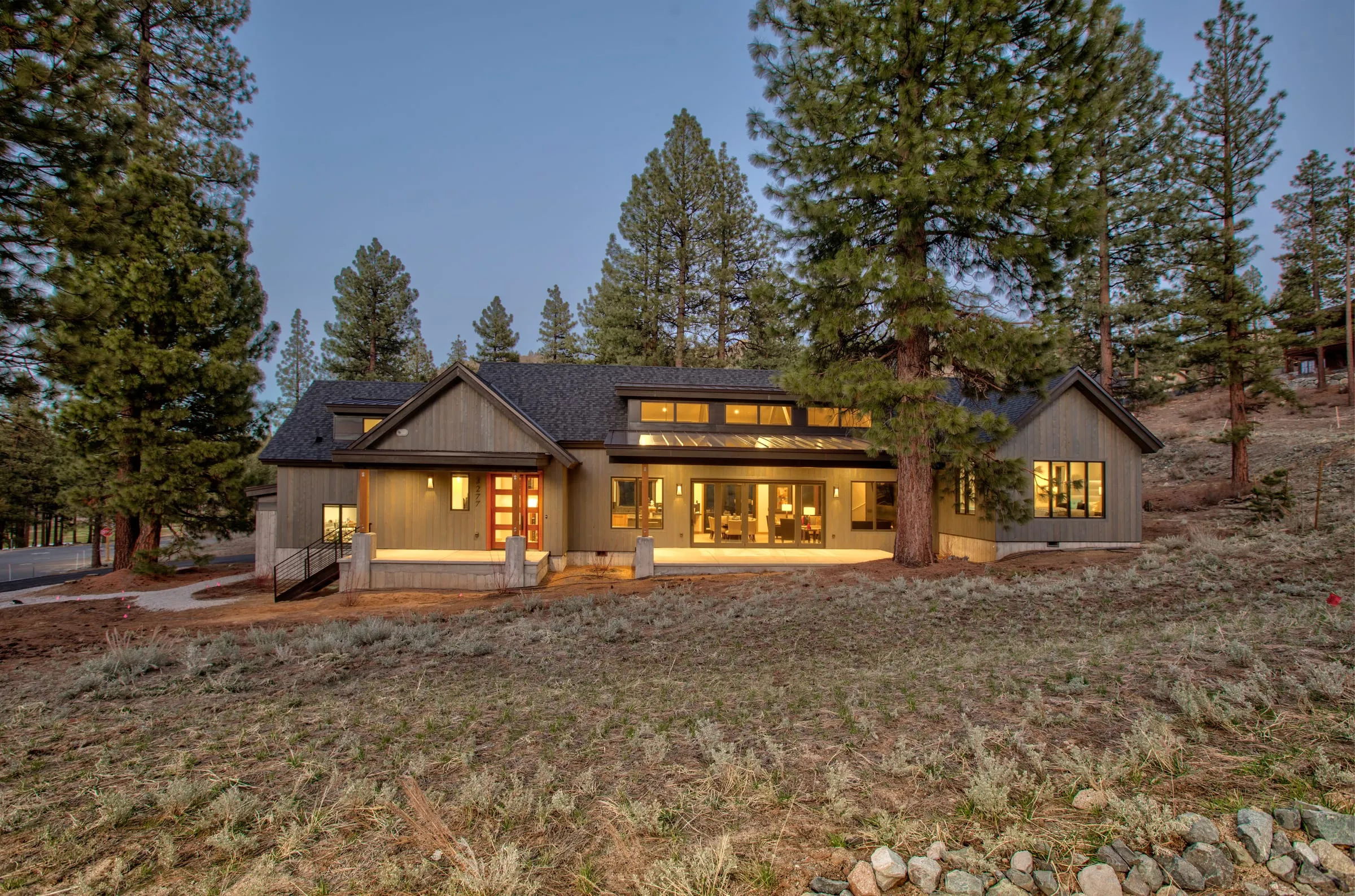 Gabled house at dusk with lit clerestory windows and glass doors along a raised terrace, set back among pines across a sagebrush slope