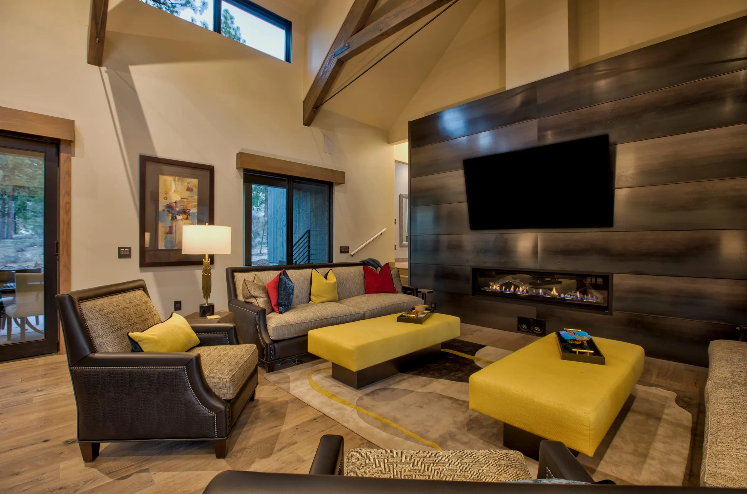 Living area with a dark wood-plank fireplace wall, a linear fireplace, yellow ottomans and a timber truss under the vaulted ceiling