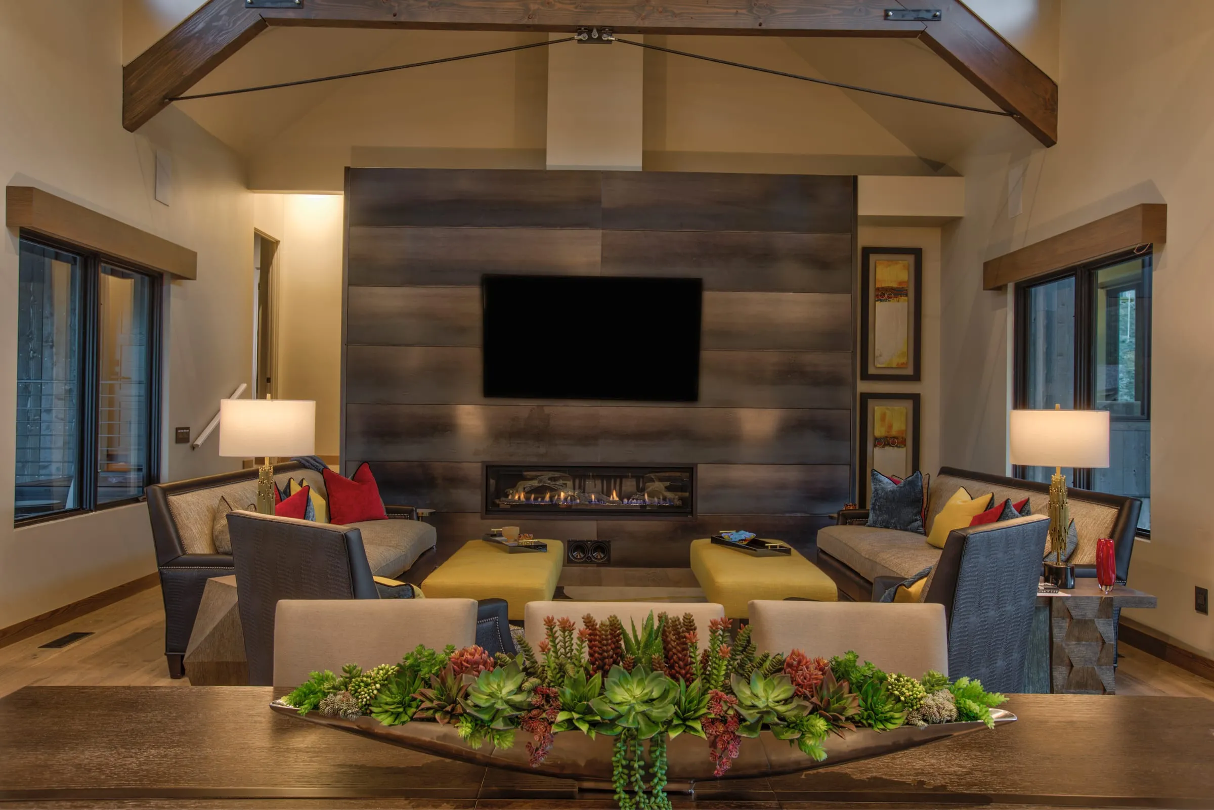 Living room seen across a succulent centerpiece on the dining table, with a plank fireplace wall, lamps and a timber truss above