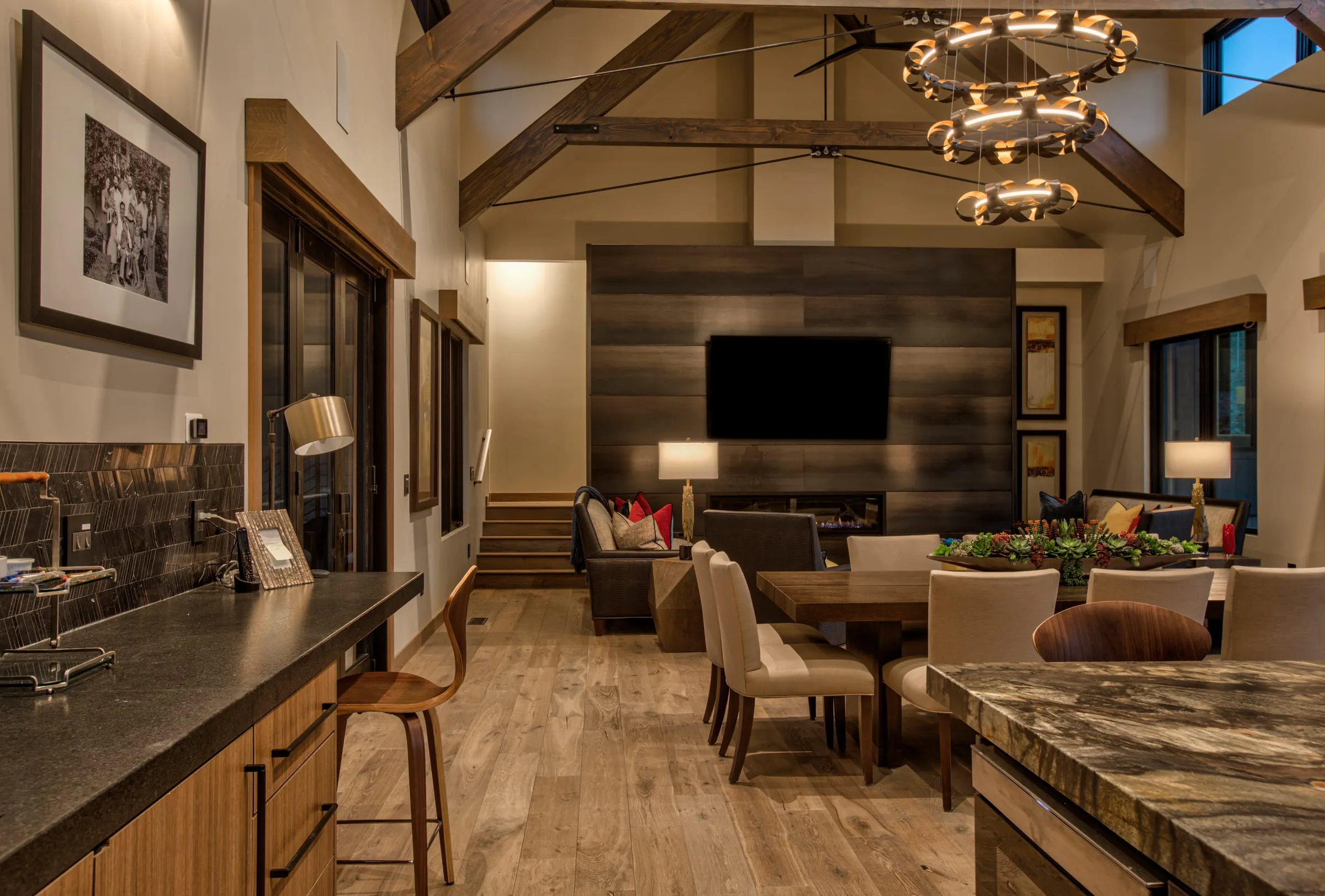 Bar counter and dining table leading to the living area, under a ring chandelier and timber trusses