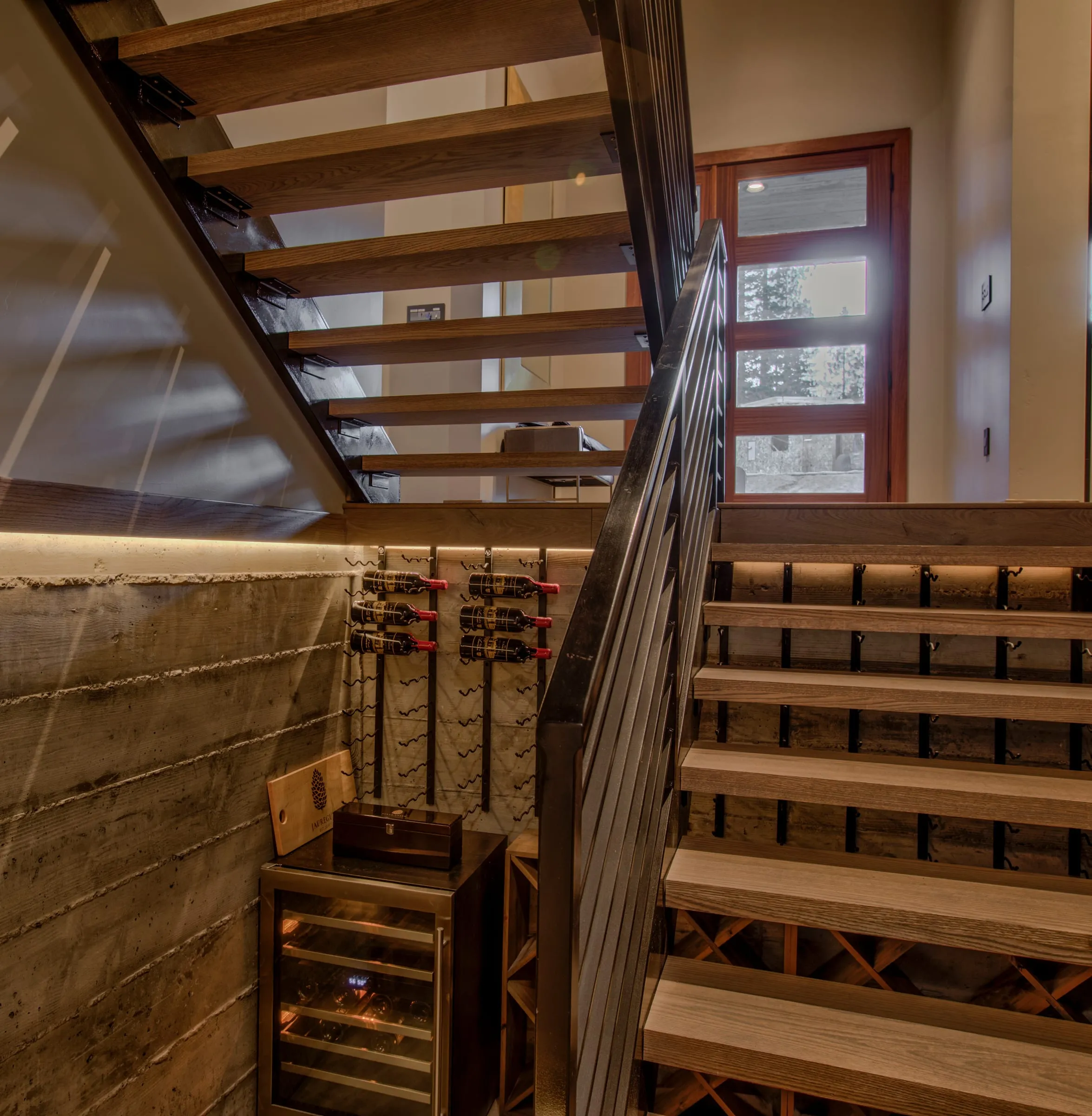 Open-tread wood stair over a board-formed concrete wall with wine racks and a wine fridge, the glass front door beyond