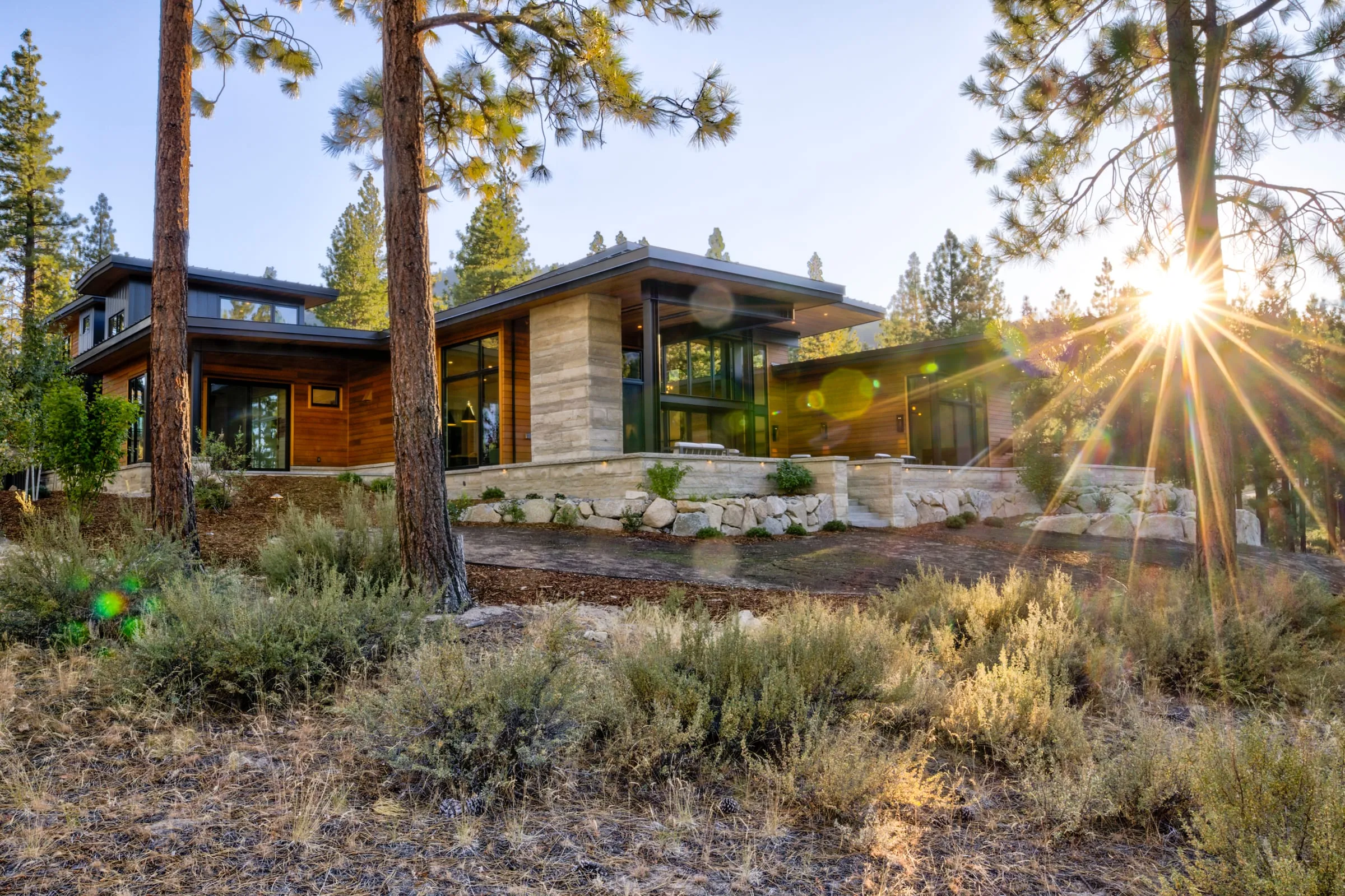 Modern home of cedar, dark siding and stacked stone on a rise of sagebrush and pines, the low sun flaring through the trees