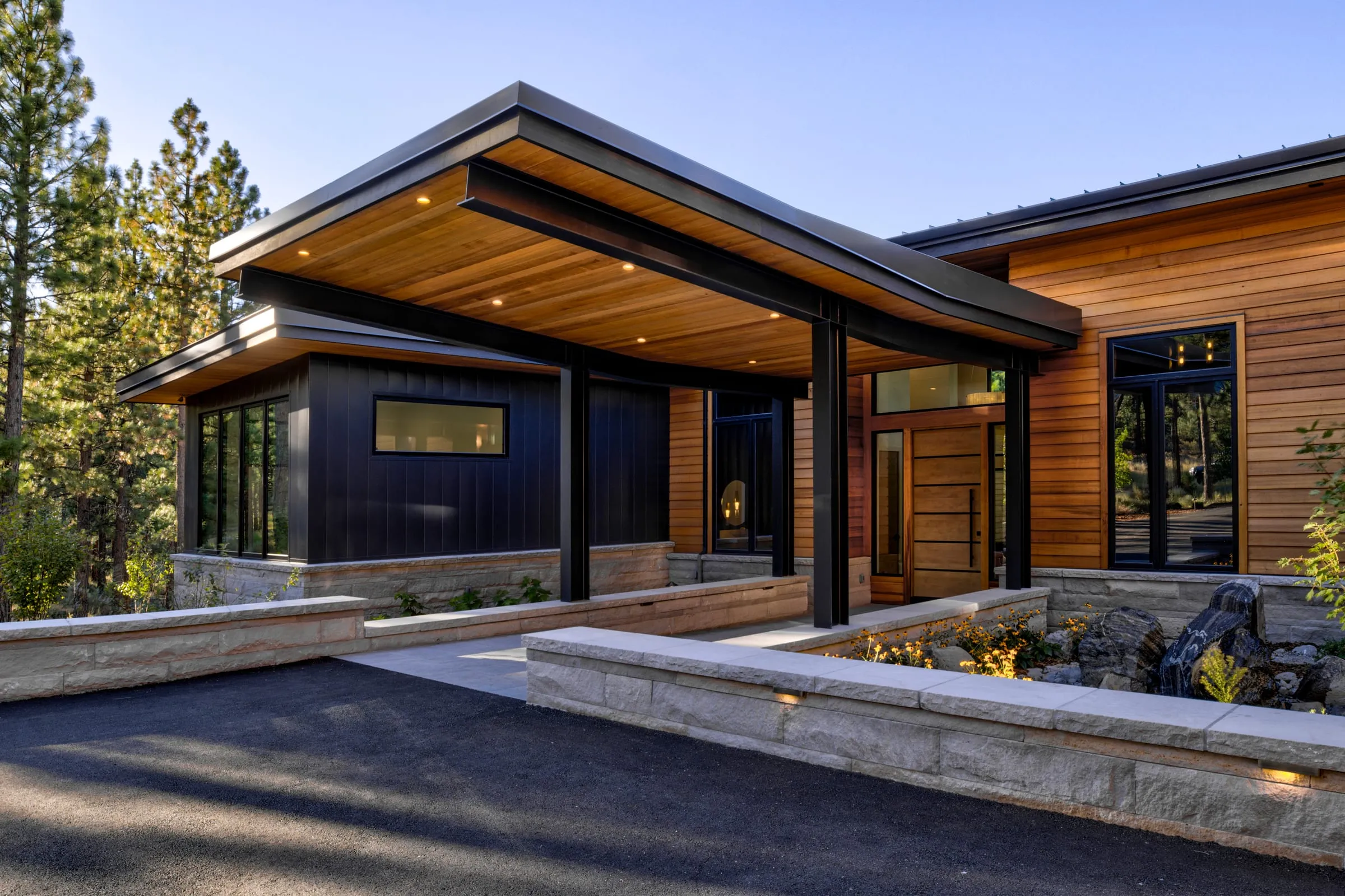 Entry court with a deep cedar-soffit canopy on black steel posts, dark vertical siding, cedar walls and lit stone planters along the drive