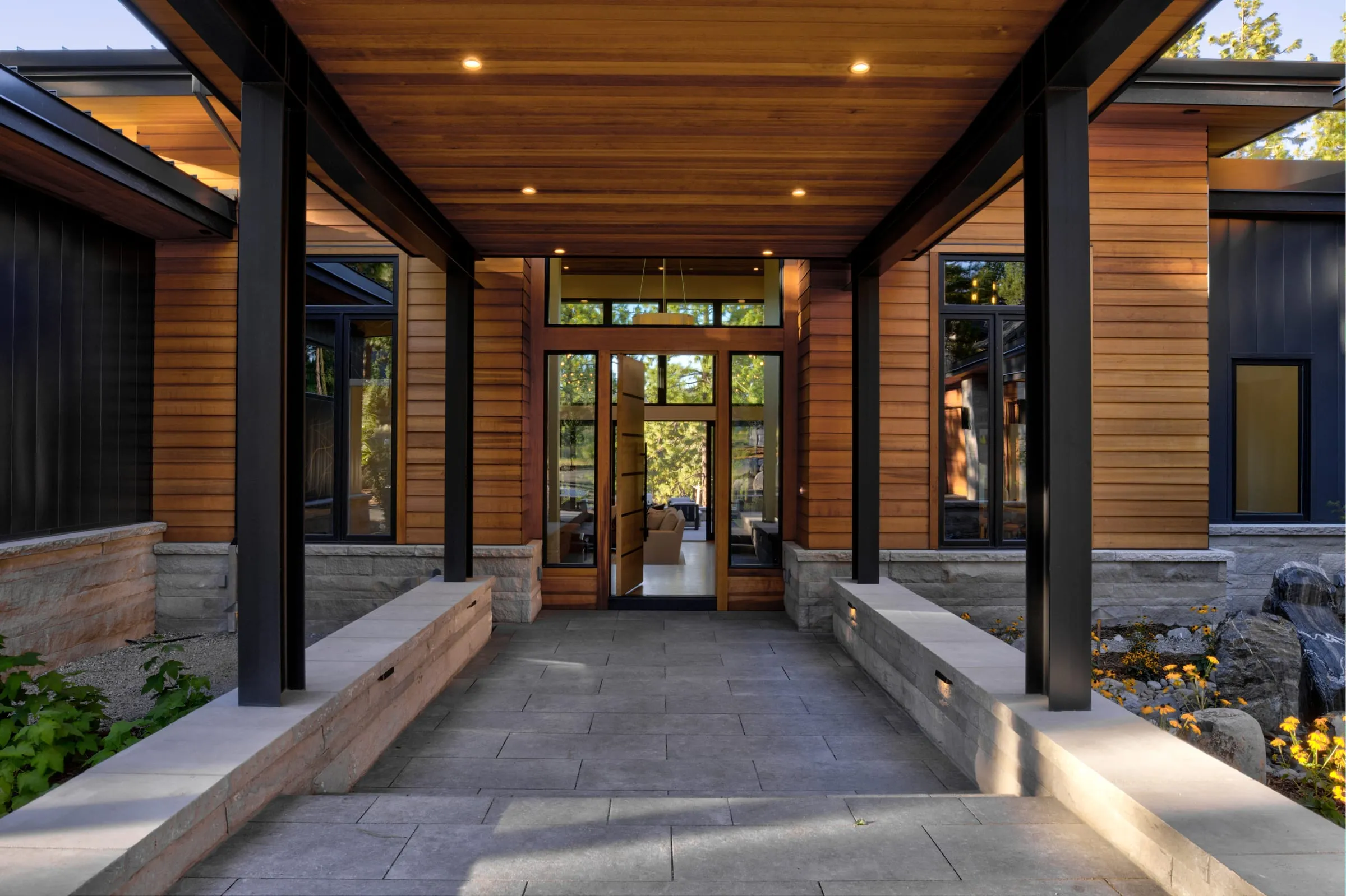 Covered entry walk between low stone walls under a cedar ceiling on black steel columns, leading to a glass front door