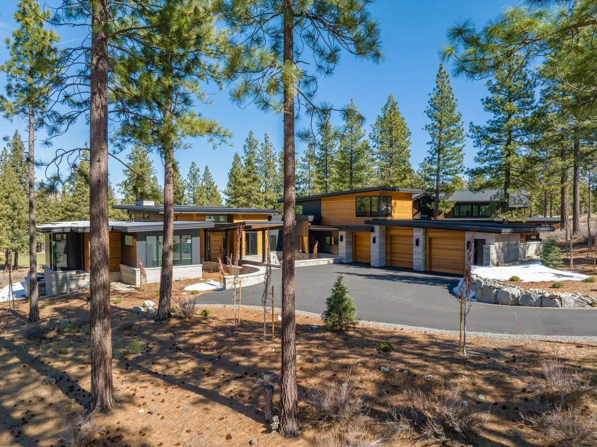 Modern home with cedar and stone wings and garage doors, seen across a curving driveway through a pine forest