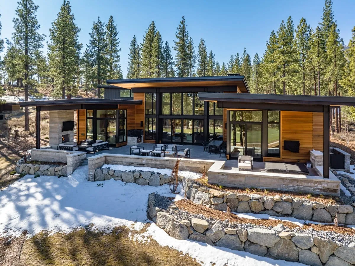 Rear of a modern home with glass walls, covered terraces and an outdoor fireplace above boulder retaining walls and melting snow