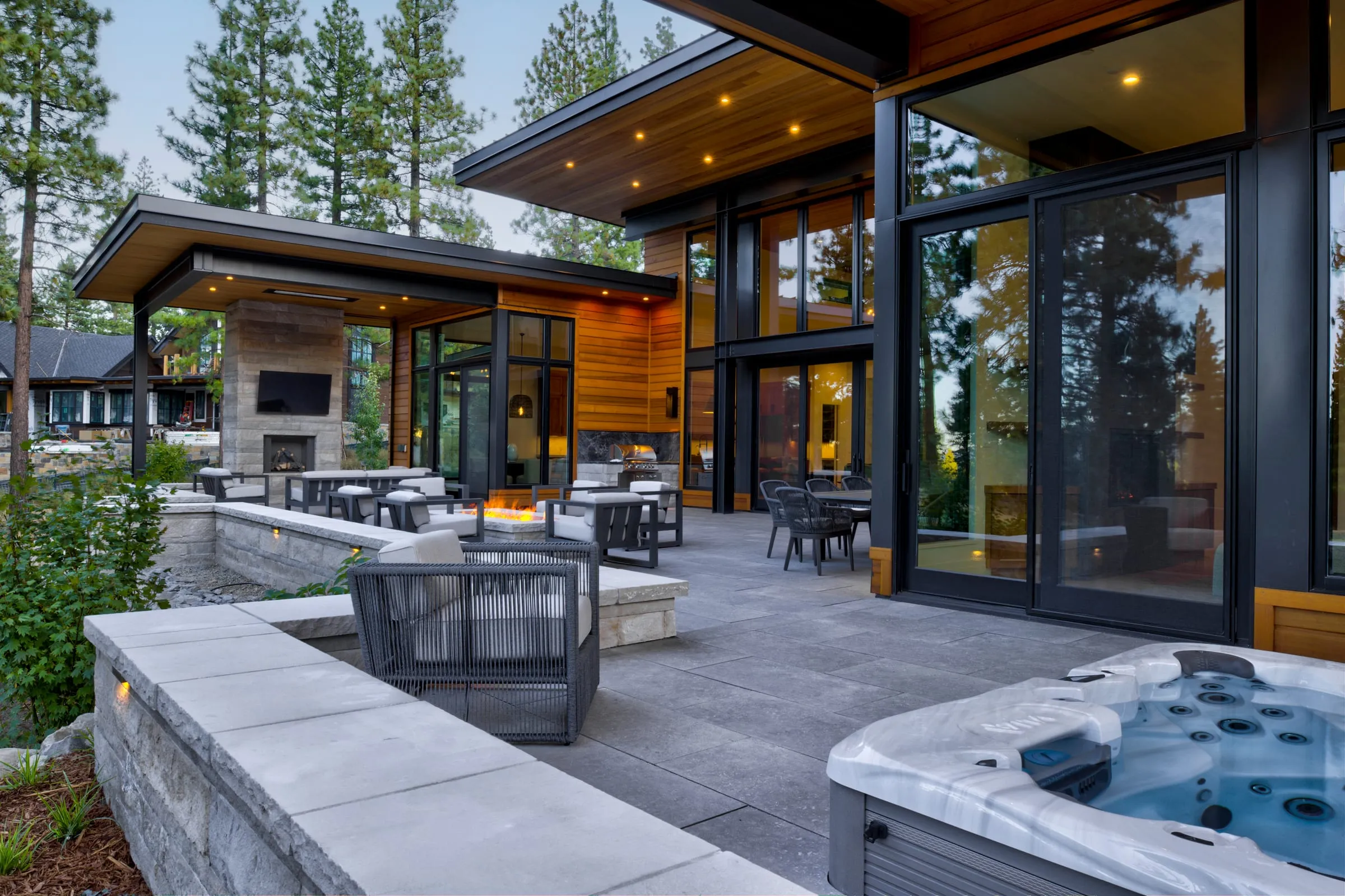Stone-paved terrace with a hot tub, lounge chairs, dining tables and an outdoor fireplace beside tall glass doors at dusk