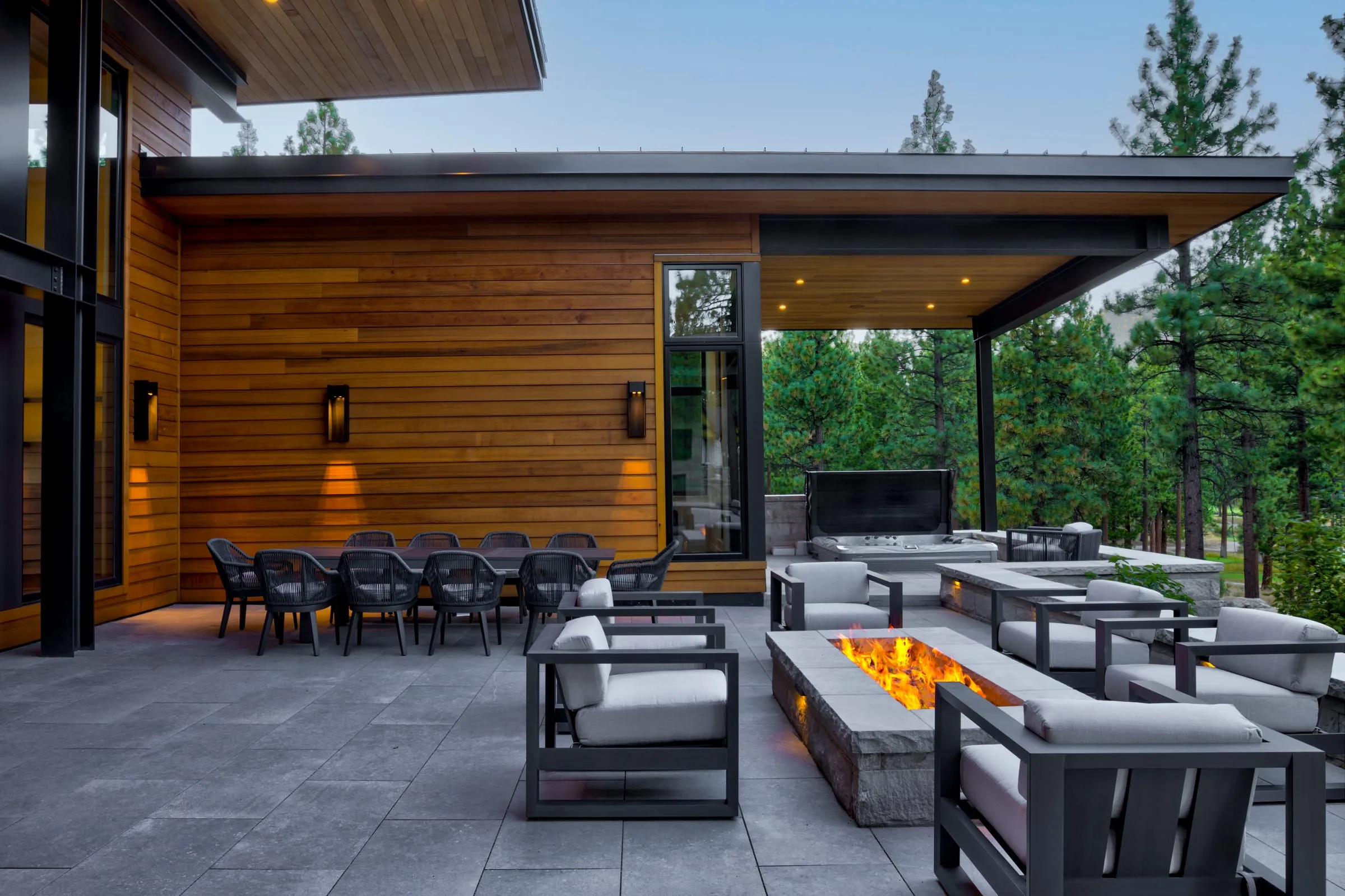Covered terrace with a long outdoor dining table against a cedar wall with lights, and lounge chairs around a fire table