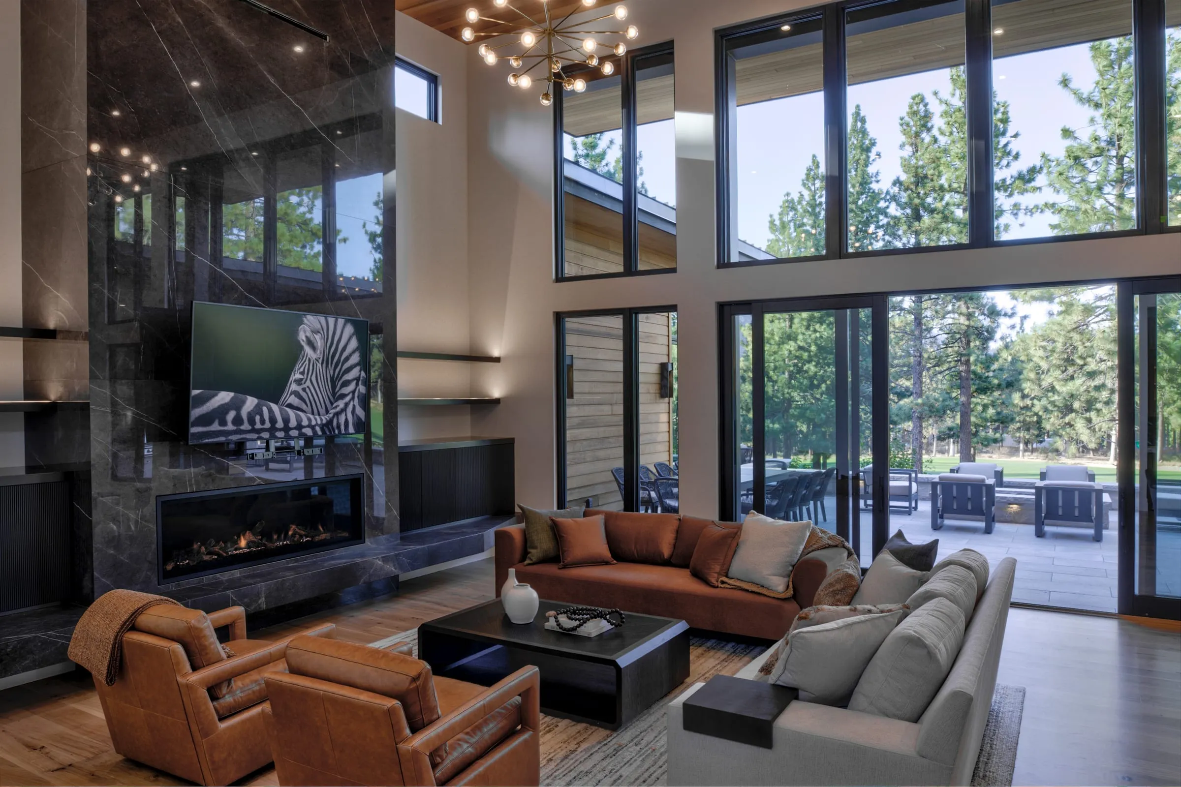 Double-height living room with a dark stone fireplace wall, leather chairs and sofas before tall windows and doors to the terrace