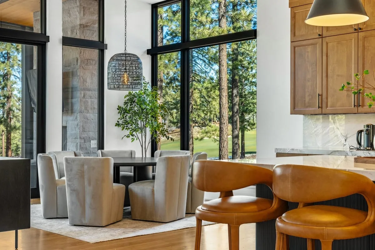 Dining area with upholstered chairs around a round table under a woven pendant, beside tall windows to the pines and a golf fairway