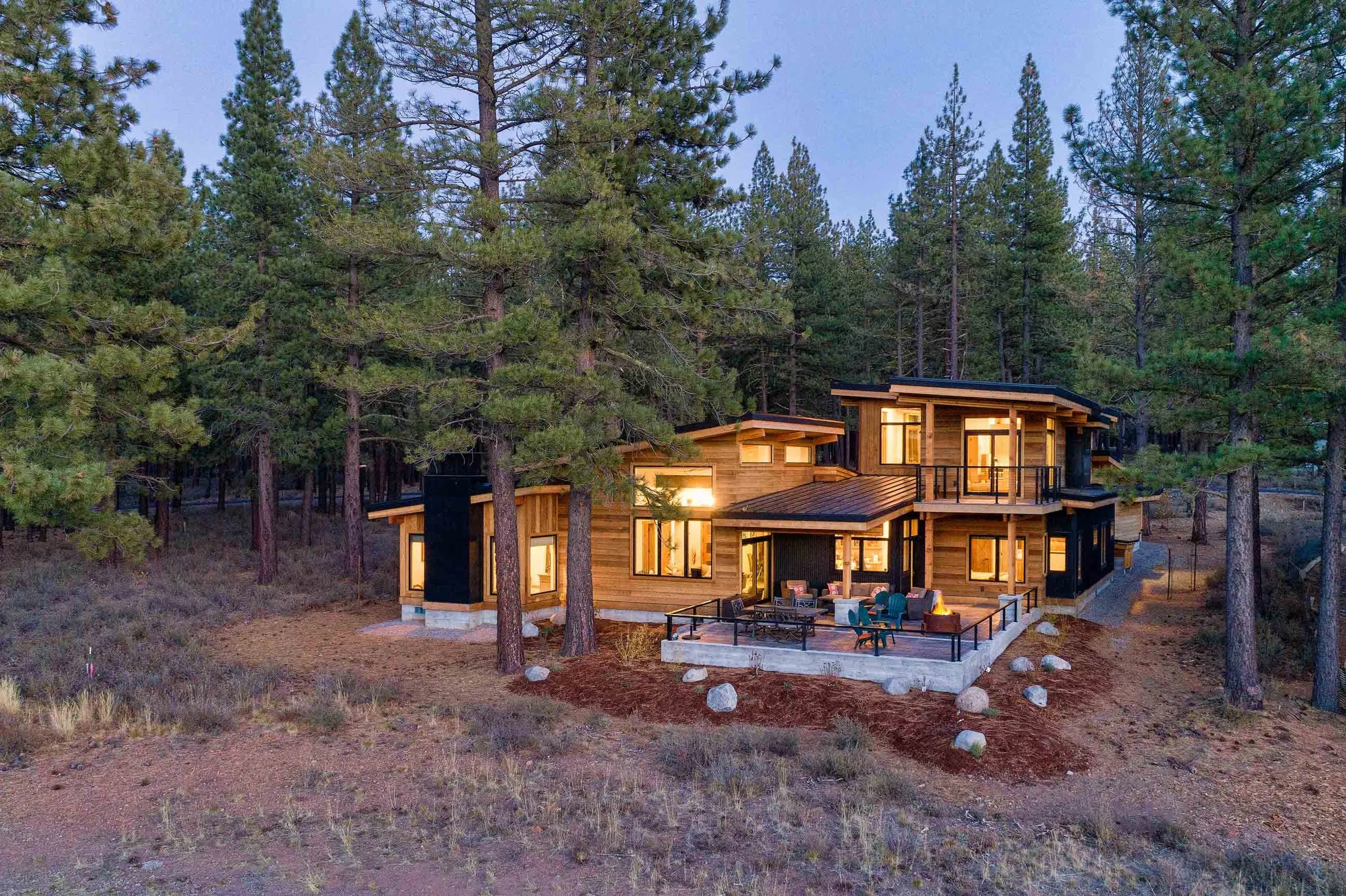Aerial view at dusk of a two-story wood and glass contemporary house with a terrace, fire pit and balcony in a pine forest