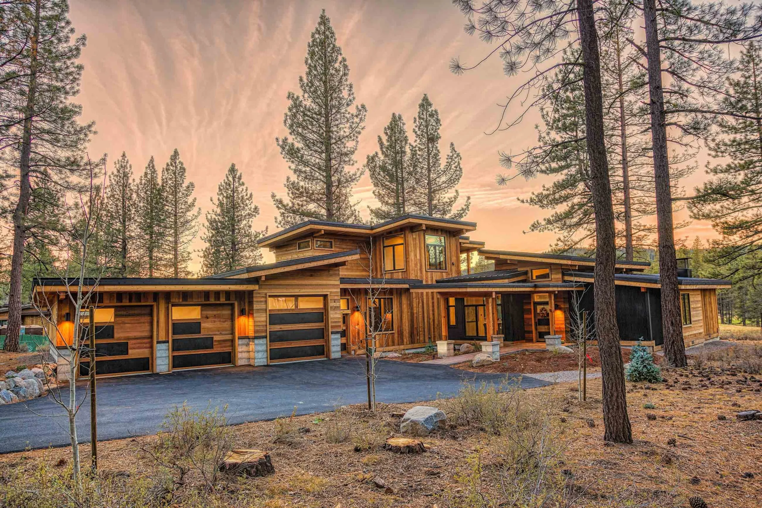 Contemporary mountain house at sunset with three garage doors, stained wood and stone, flat and shed roofs, pines and driveway