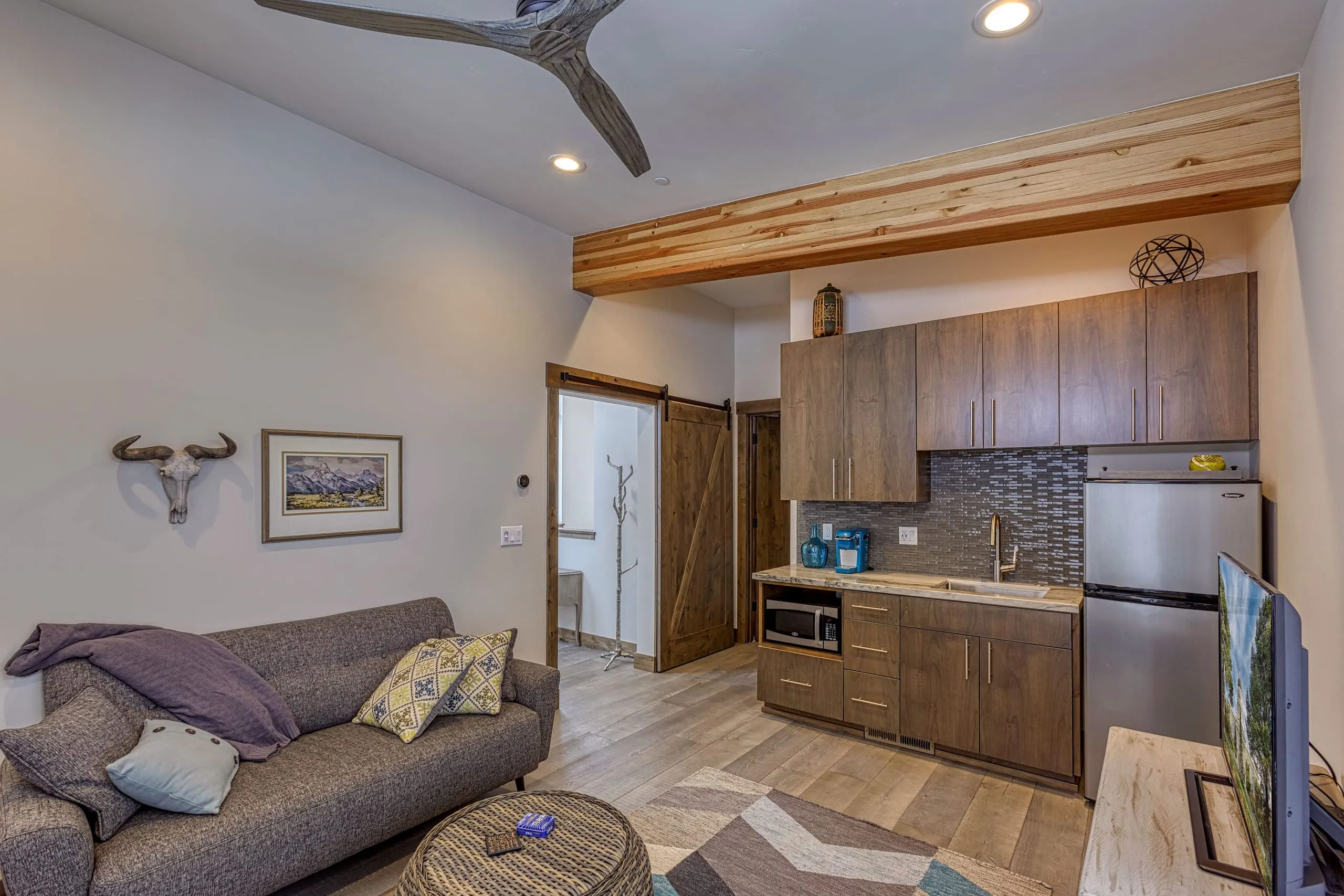 Small guest suite interior with a gray sofa, kitchenette with brown cabinets, wood beam over the counter and a barn door
