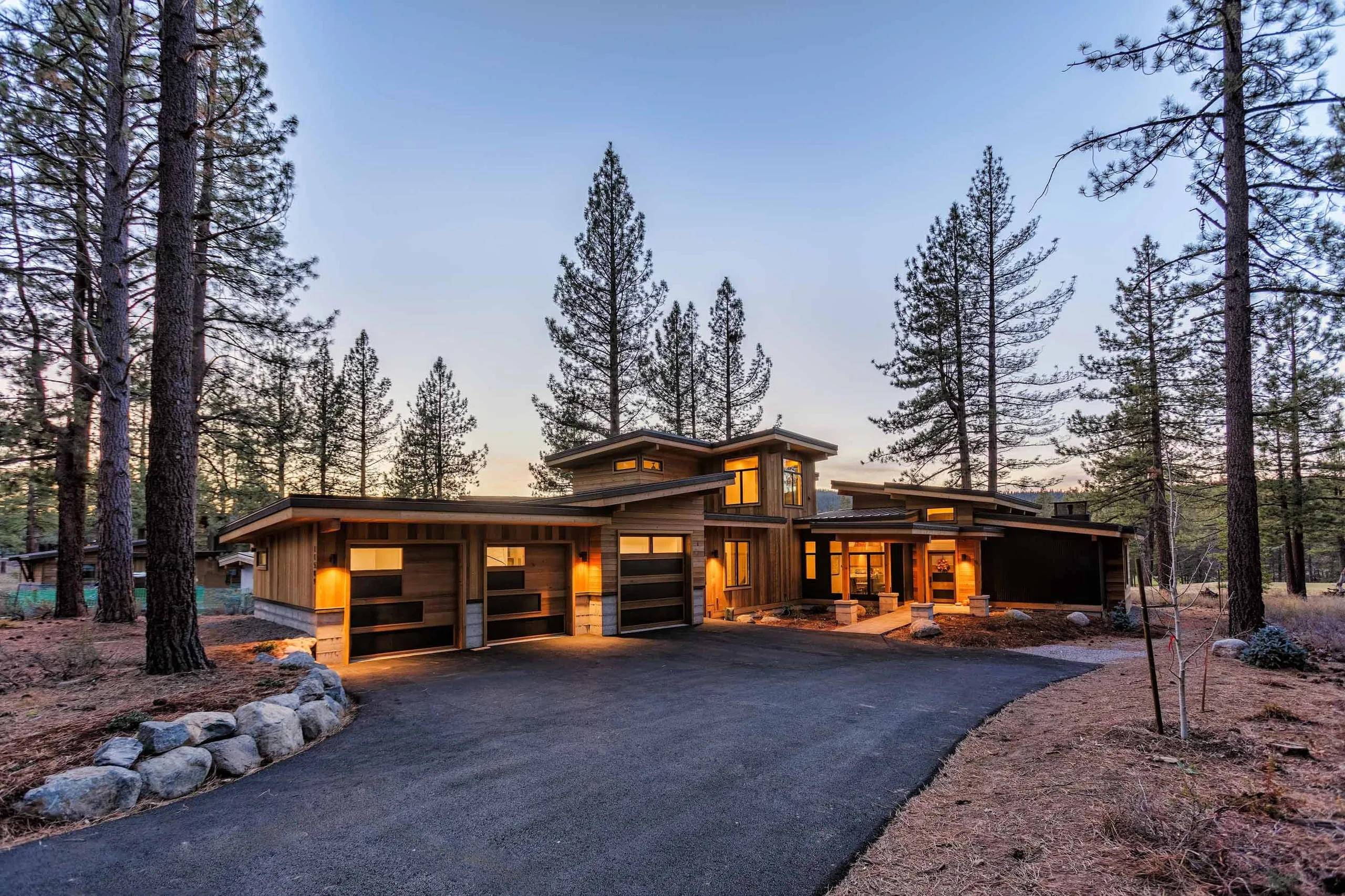 Contemporary house at dusk with lit three-car garage, wood siding and stone accents, asphalt driveway and tall pines