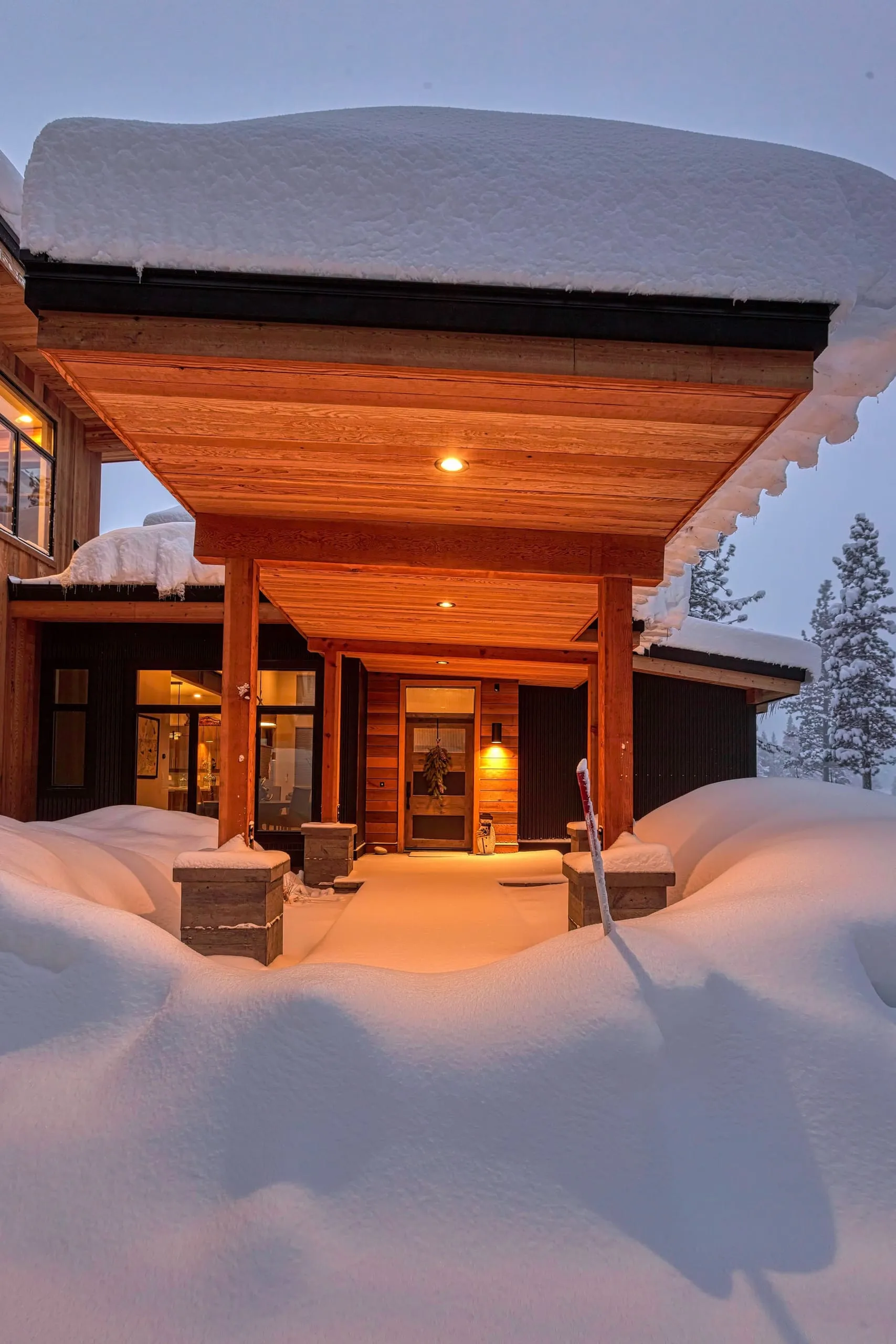 Snow-covered entry canopy with wood soffit on timber posts, stone pier bases and lit front door
