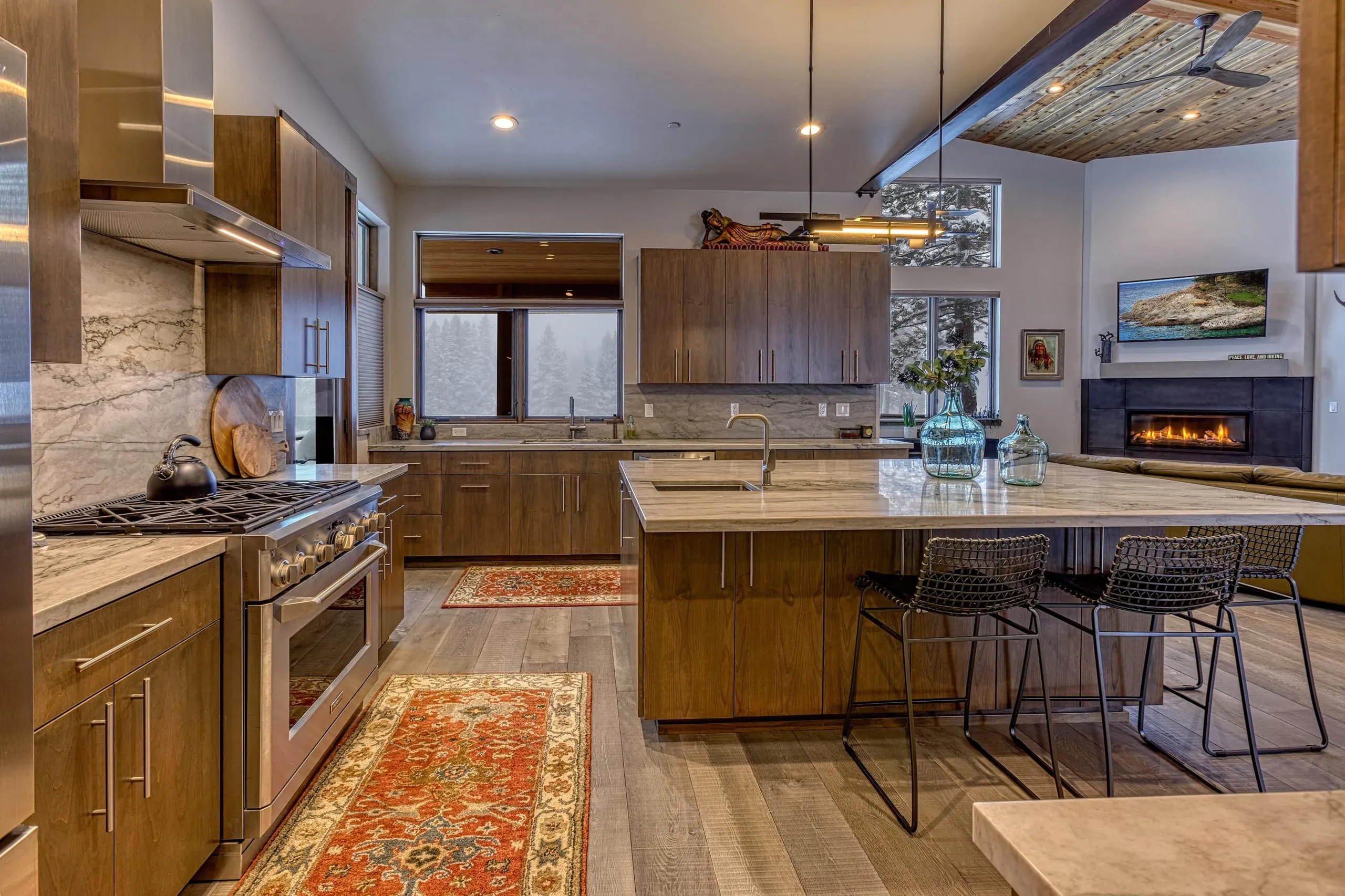Open kitchen with gas range, long stone island and stools, looking toward a fireplace and wood-ceilinged living area