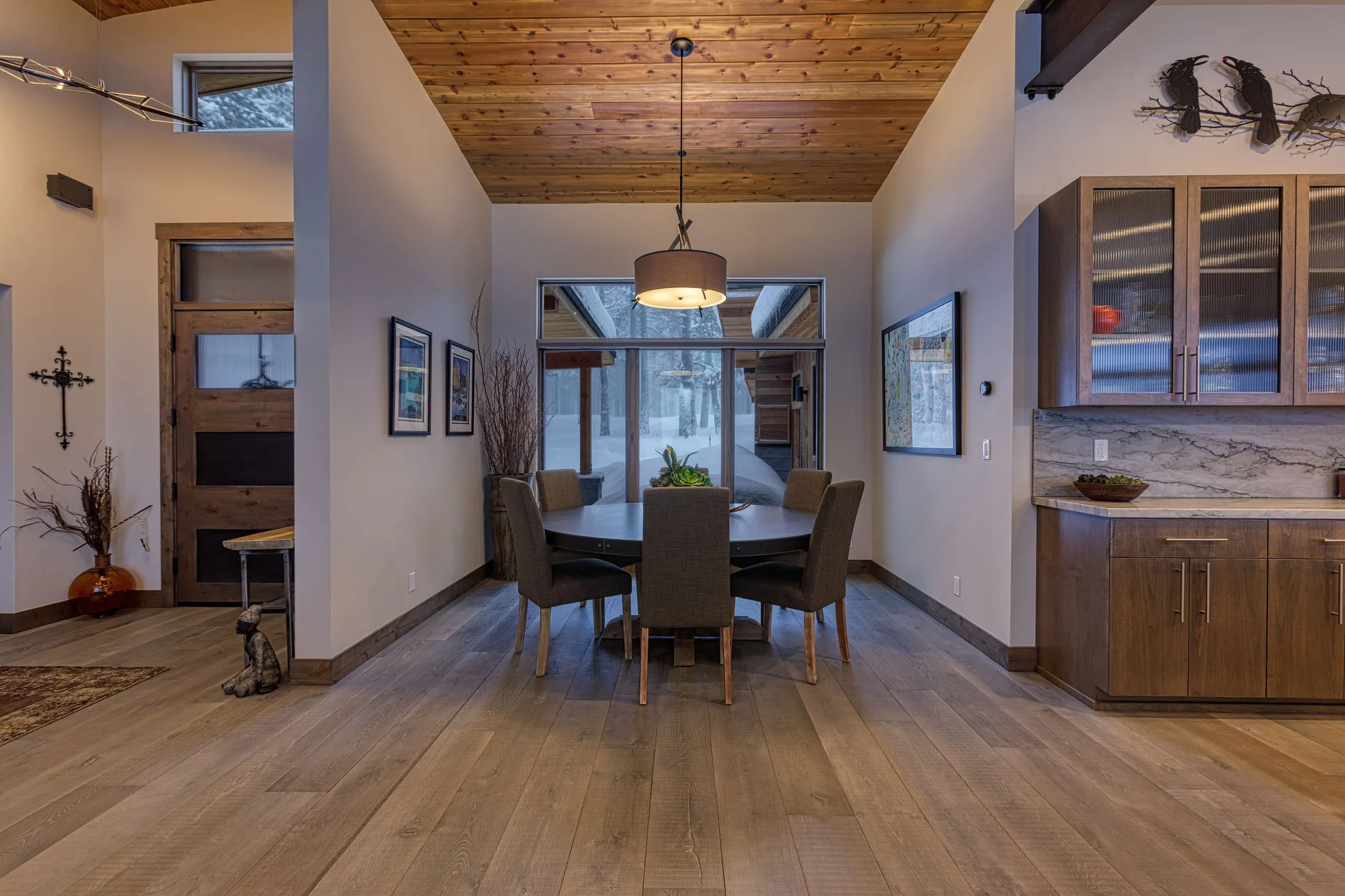 Dining area with round table and a wood ceiling, facing a large window, with kitchen cabinets at right
