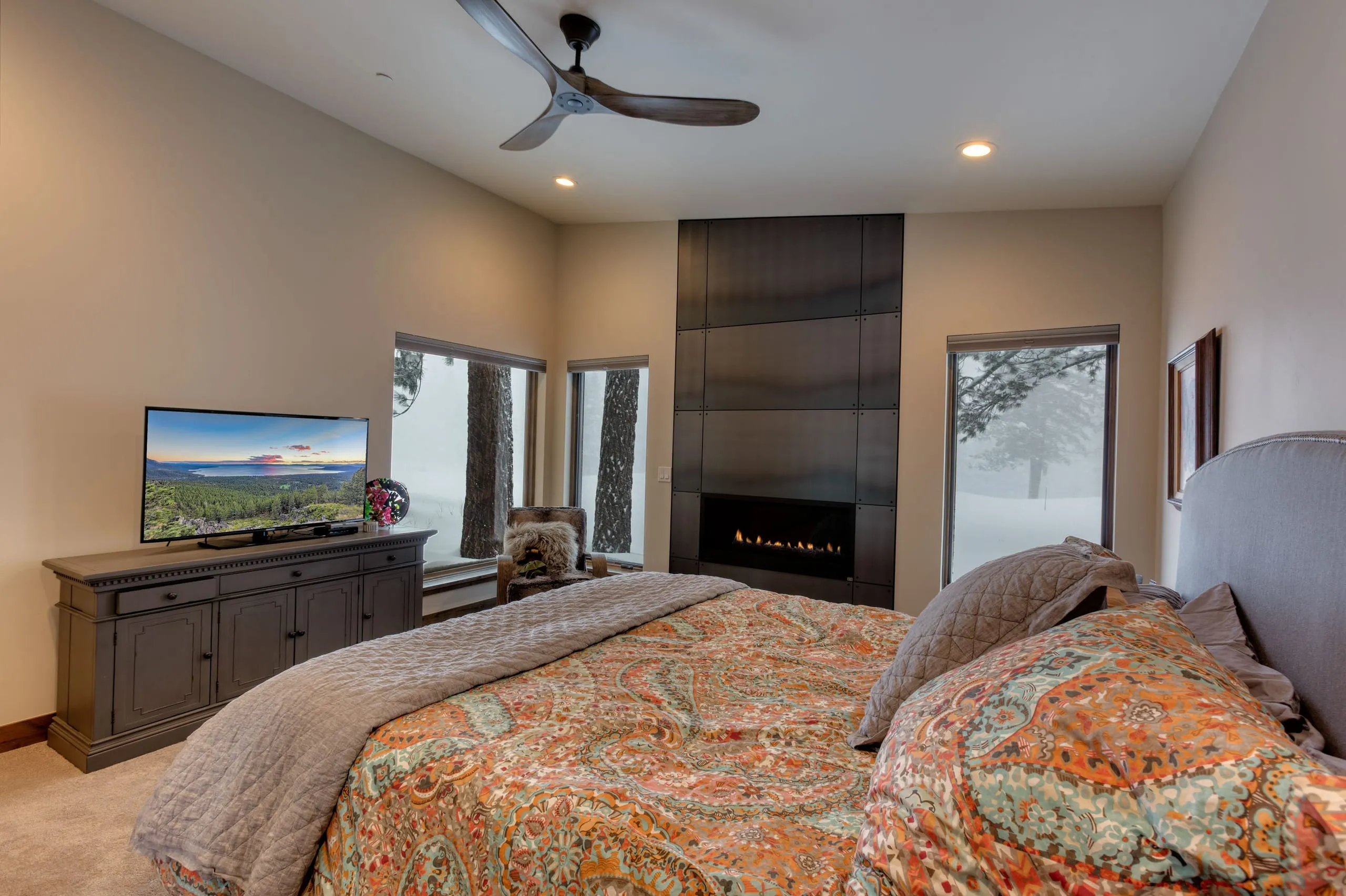 Bedroom with a dark tiled gas fireplace wall, ceiling fan, windows onto snowy pines and a paisley bedspread