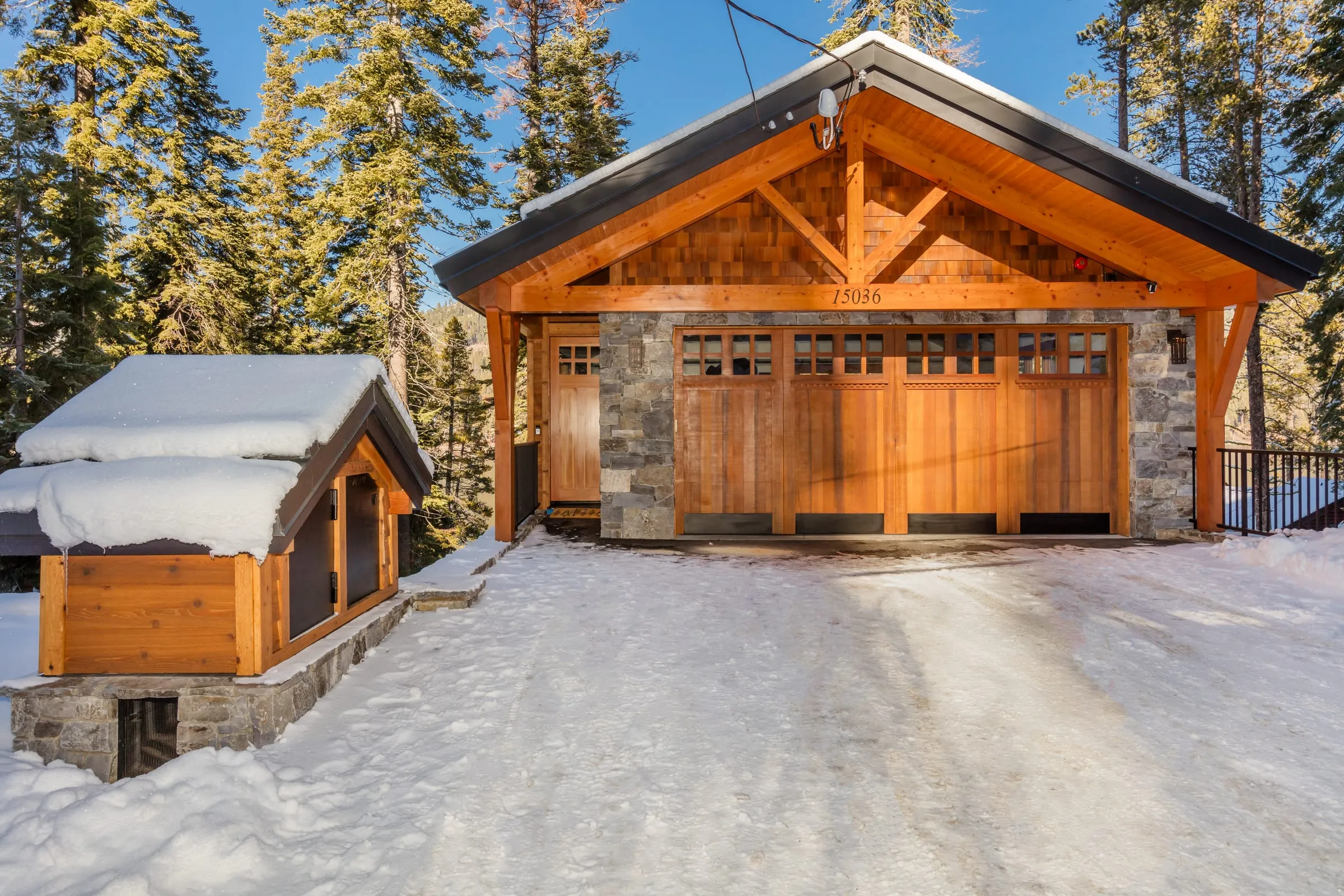 Front of a garage building in the snow, with a timber-trussed gable, shingled gable end, wood carriage doors and stone walls