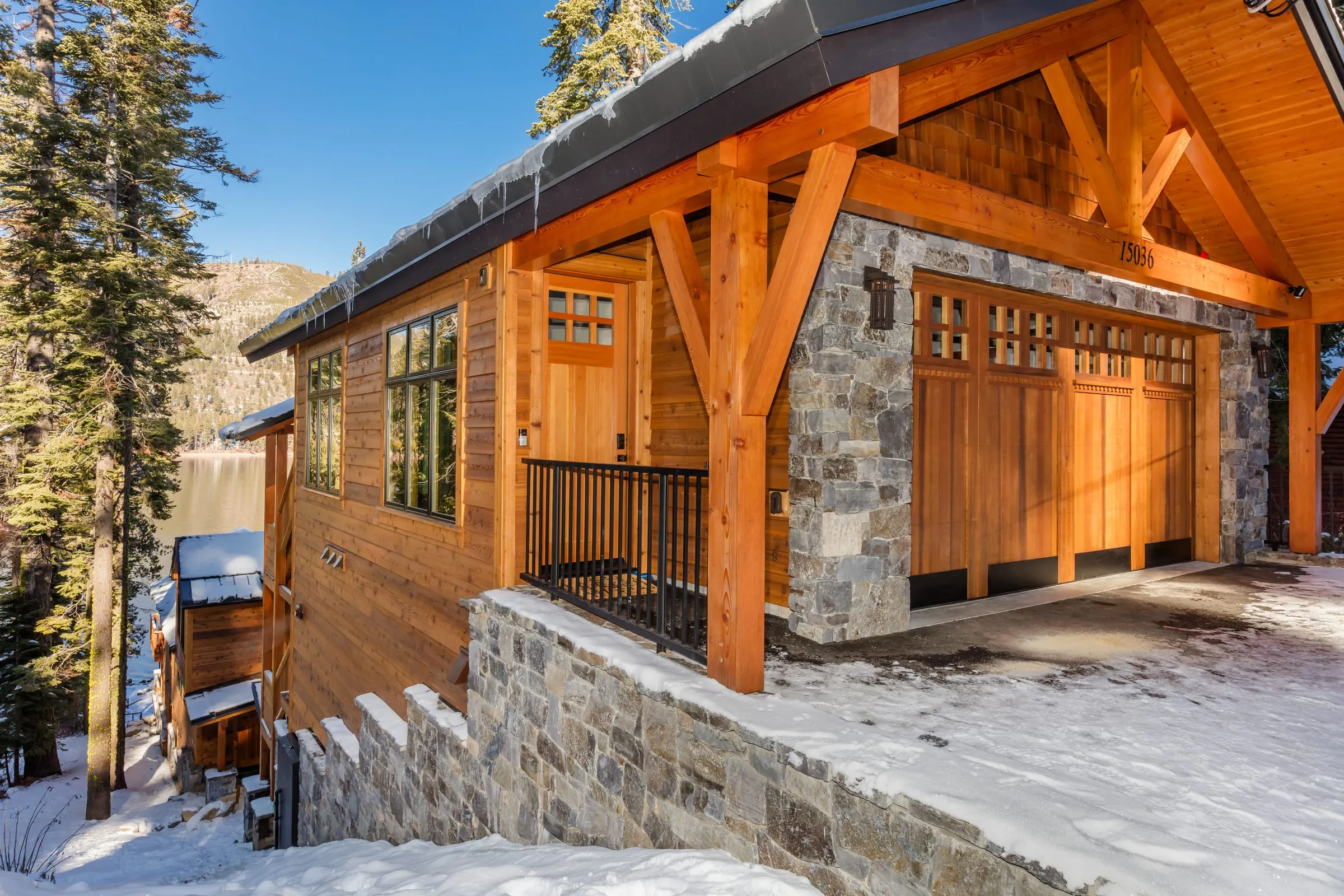 Covered entry under a timber-framed gable with a wood door, black railing, cedar siding and a stone wall, the lake beyond