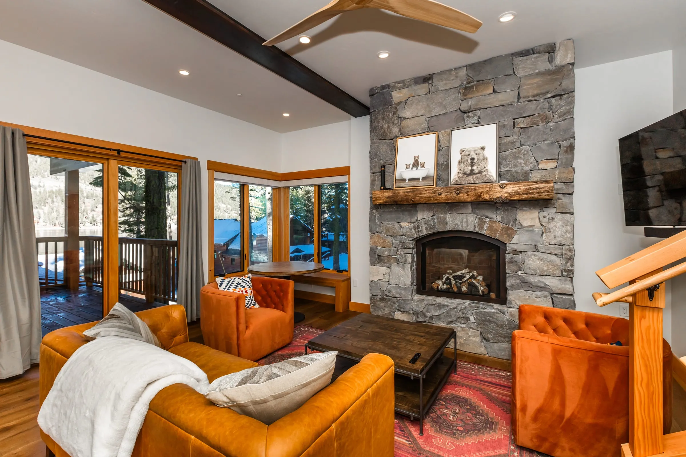 Living room with a stacked stone fireplace and log mantel, orange chairs, a leather sofa and glass doors to a deck over the lake