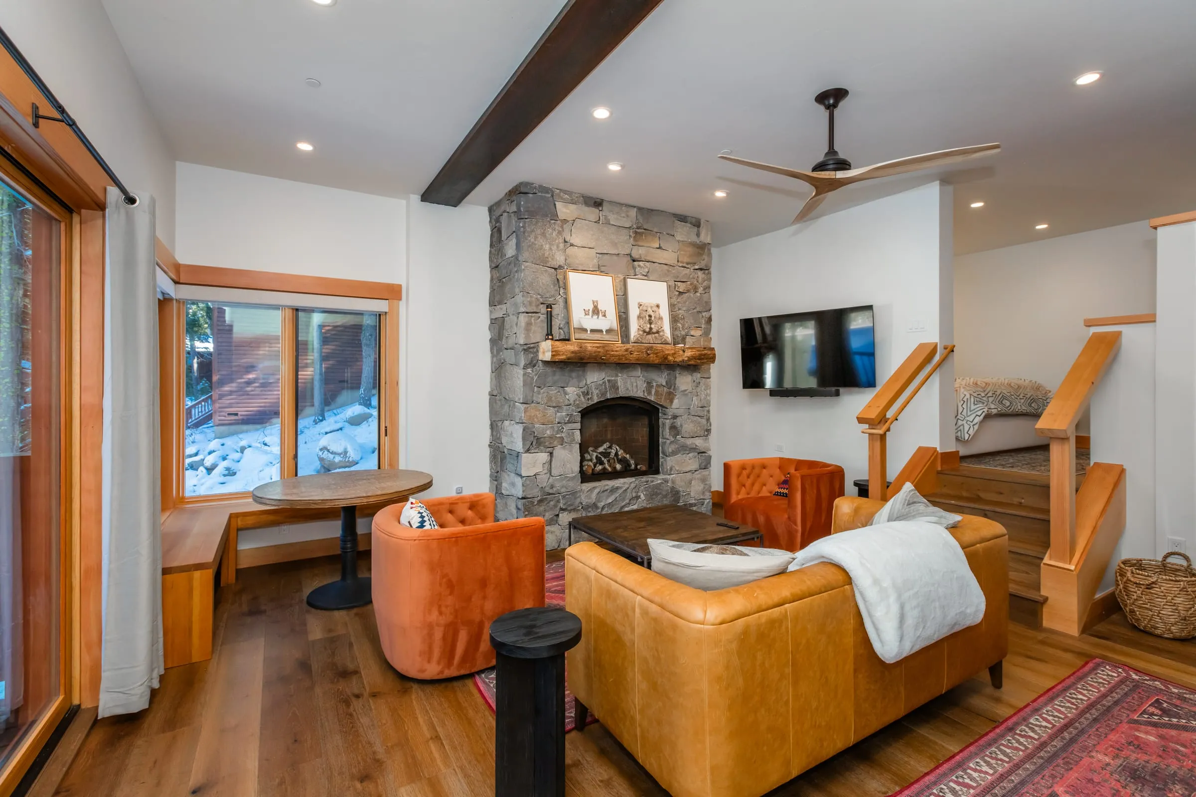 Living room with a stone fireplace, a window seat with a small table, a leather sofa and wood stairs leading up
