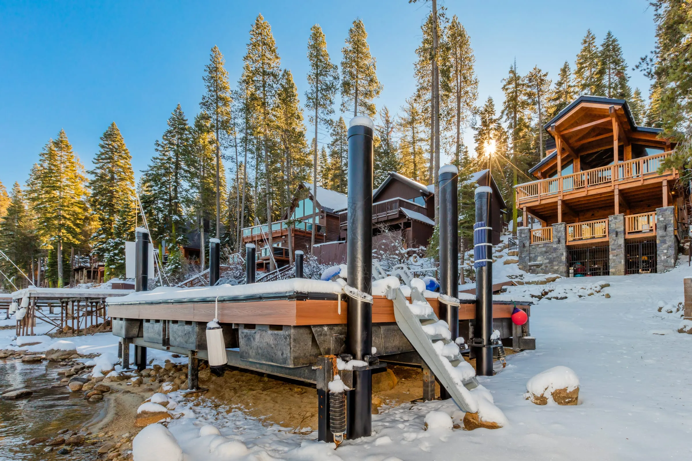 Snow-covered dock on the lakeshore with the timber-framed house and its decks among tall pines behind, the sun low through the trees