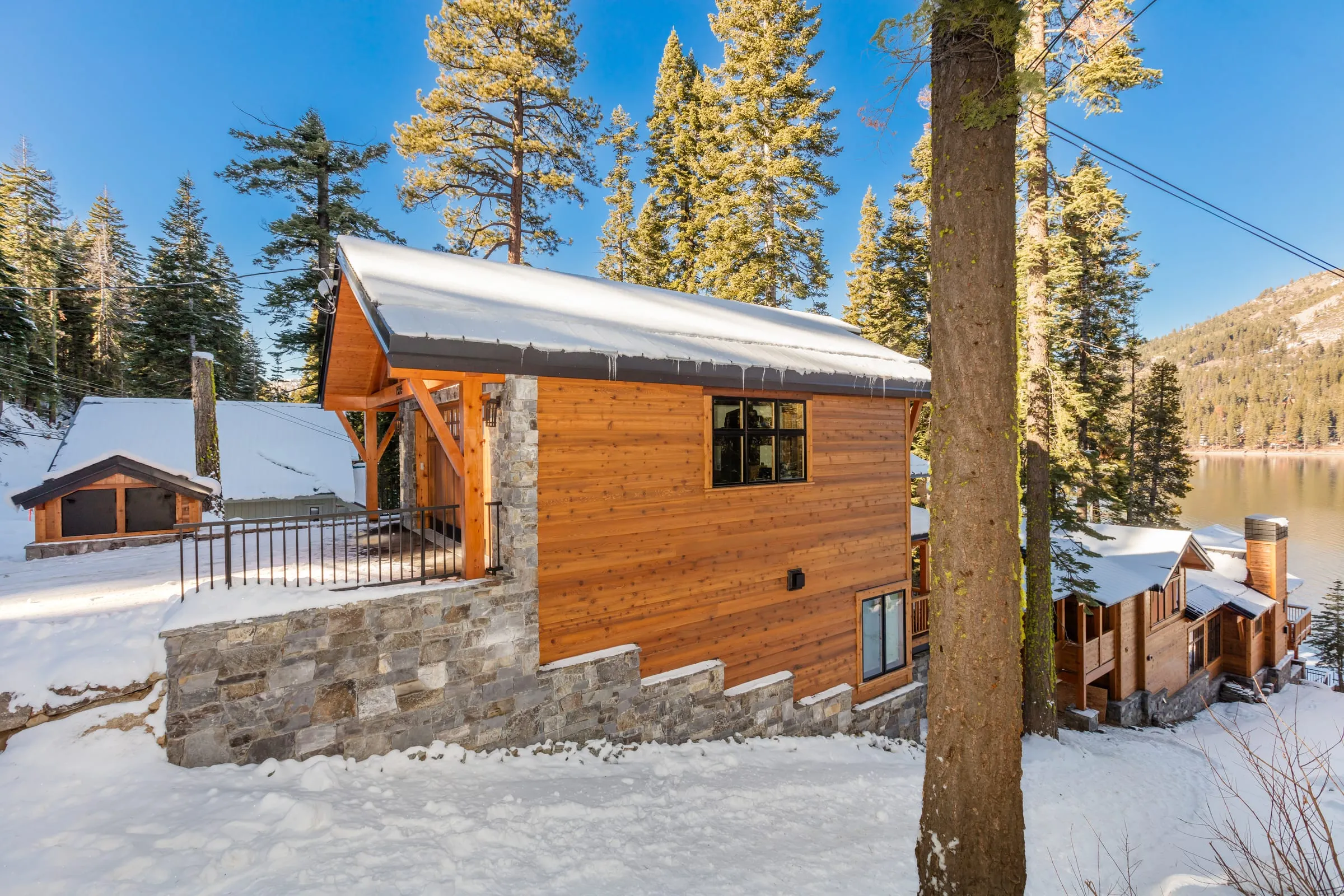 Wood-sided building with a timber gable and a stacked stone base in the snow, with the lake and a mountain ridge beyond