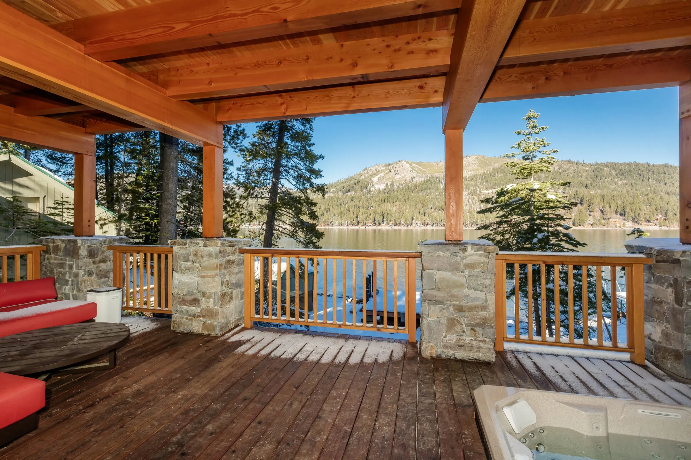 Covered deck with timber beams, stone piers and wood railings, a hot tub and red cushions, looking out over the lake to the mountains