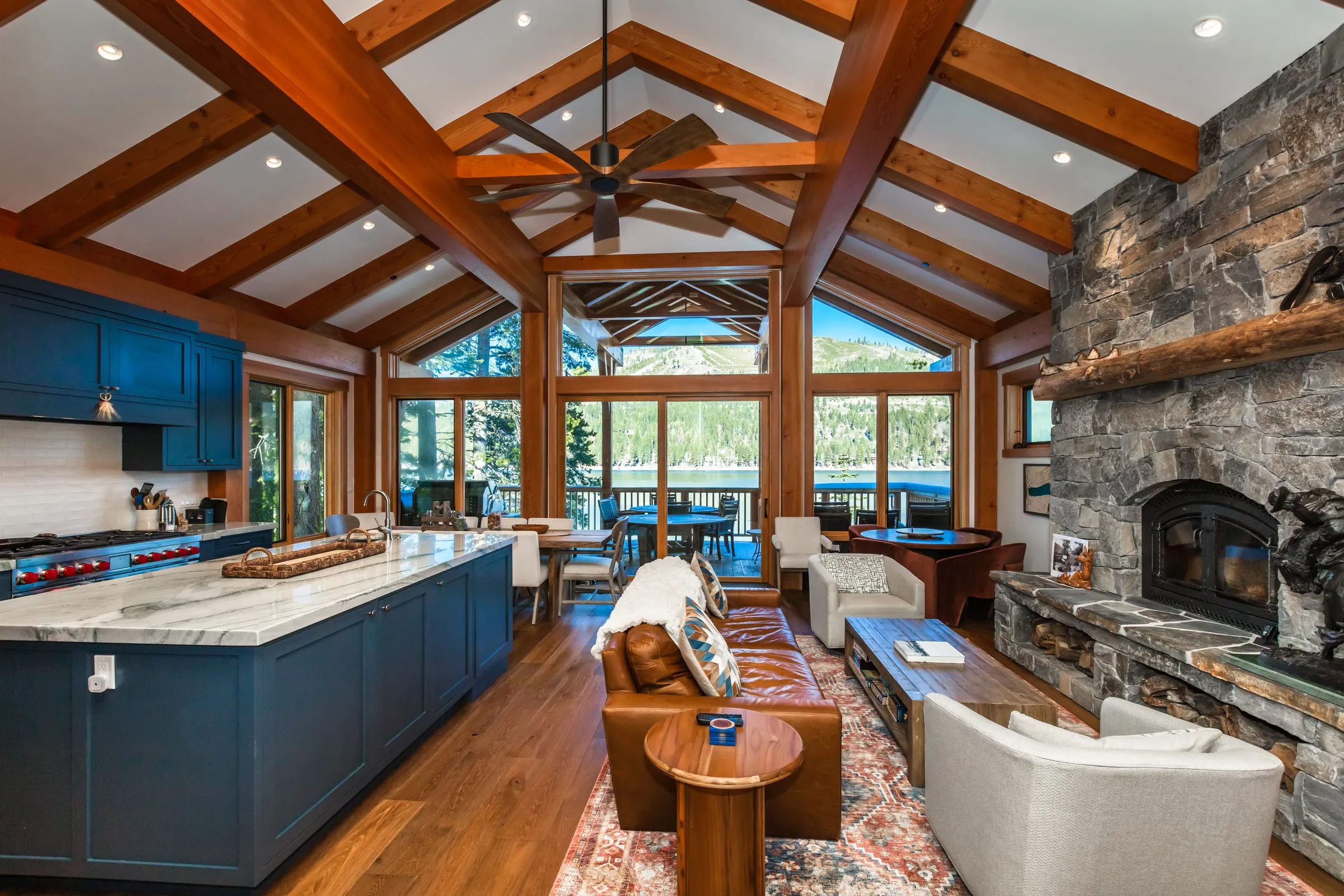 Open kitchen and living room under exposed timber trusses, with a blue island, a stone fireplace and gable windows to the lake