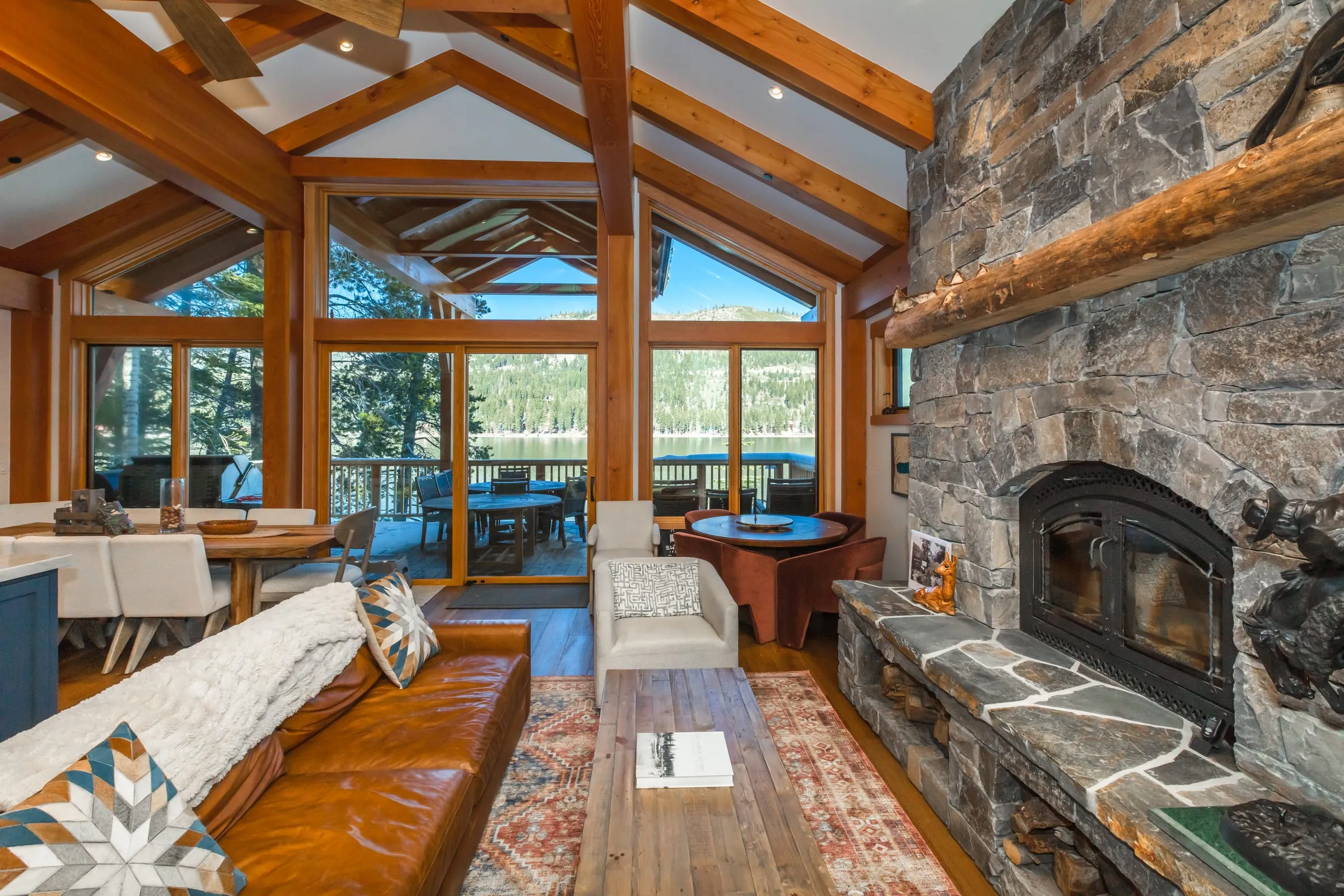 Living room with a tall stacked stone fireplace and log mantel, a leather sofa and windows and doors to a lakeside deck