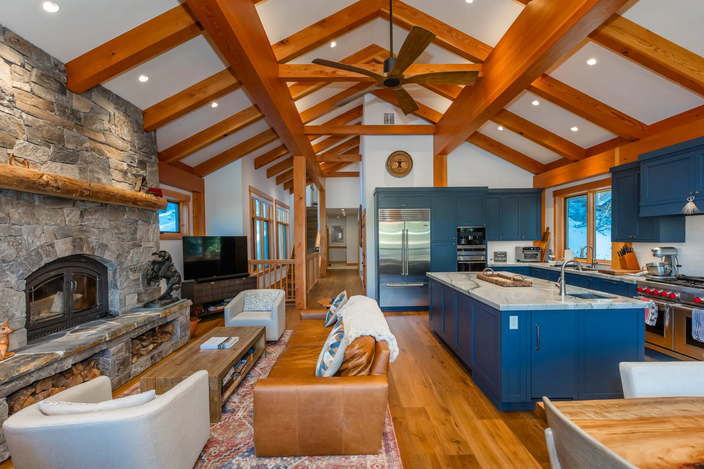 Vaulted great room with timber trusses, a stone fireplace, a leather sofa and the blue-cabinet kitchen and island beyond