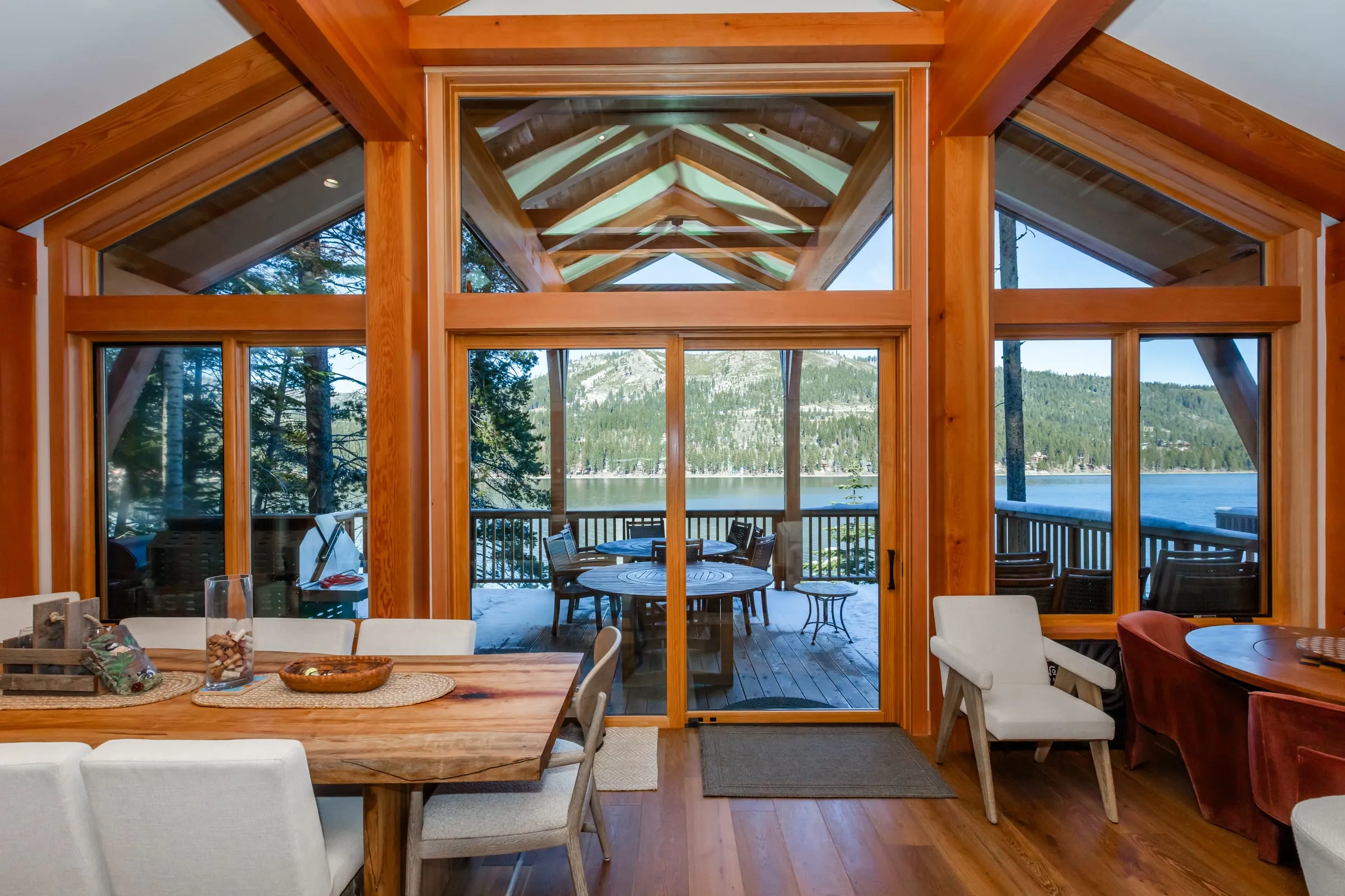 Dining table beneath a timber-framed gable wall of windows, with glass doors to a deck and the lake and mountains beyond