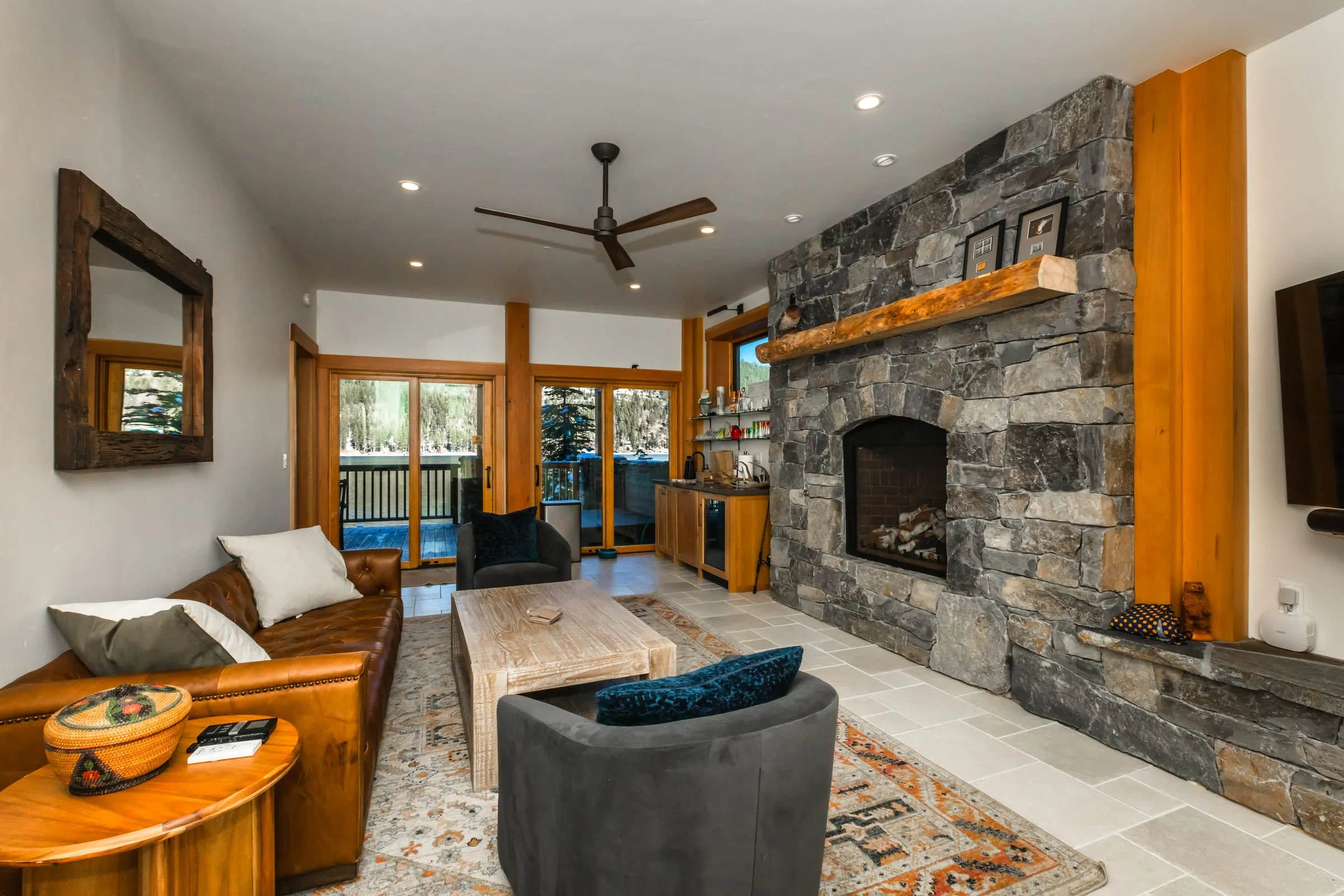 Living room with a stacked stone fireplace and timber mantel, a leather sofa, a stone floor and glass doors to a deck