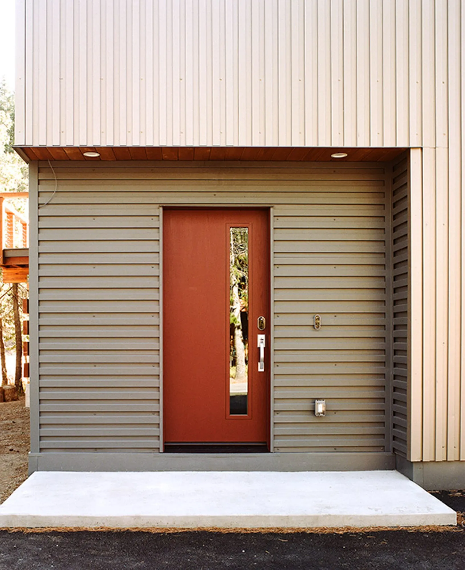 Red front door set in horizontal gray siding beneath vertical metal cladding, with a concrete stoop