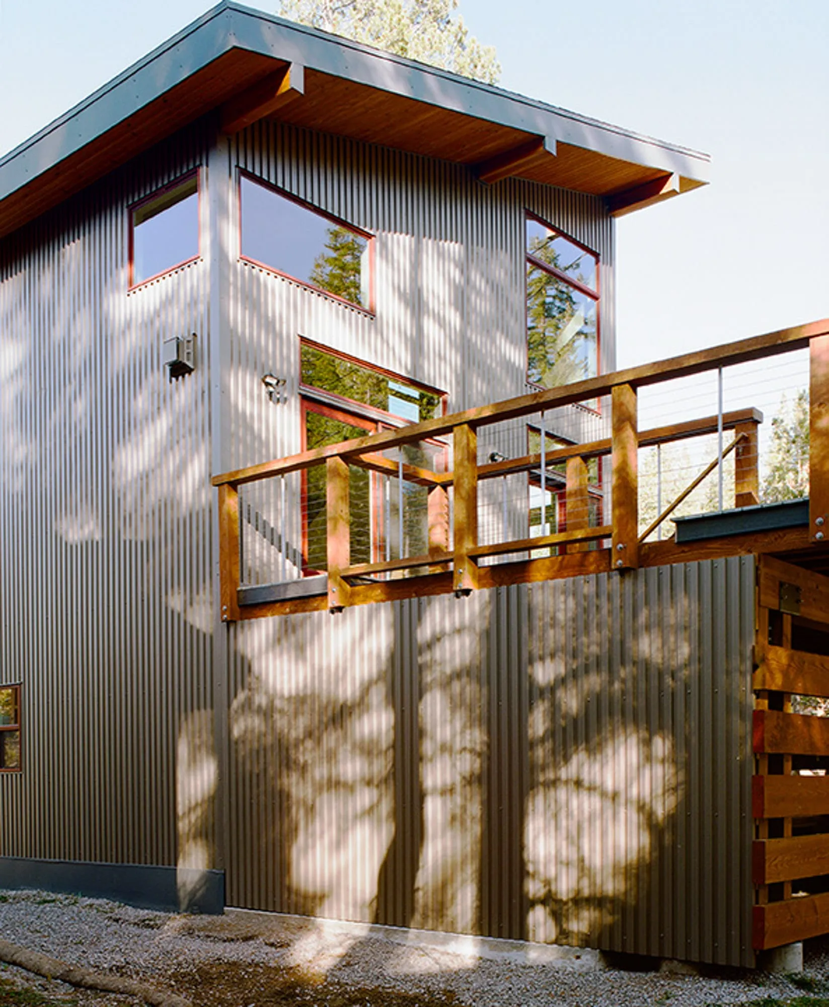 Corner of a corrugated metal house with a shed roof, wood deck with cable railing and tree shadows
